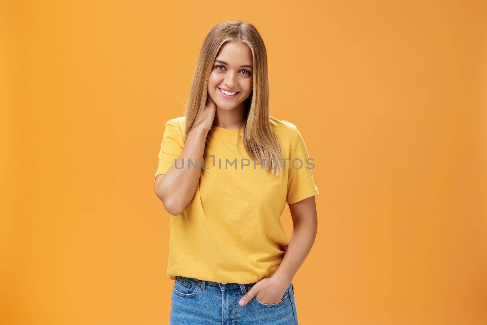 Portrait of shy and timid feminine girl with tan and straight fait hair rubbing neck and smiling sensually with happy carefree expression holding hand in pocket posing against orange background by Benzoix