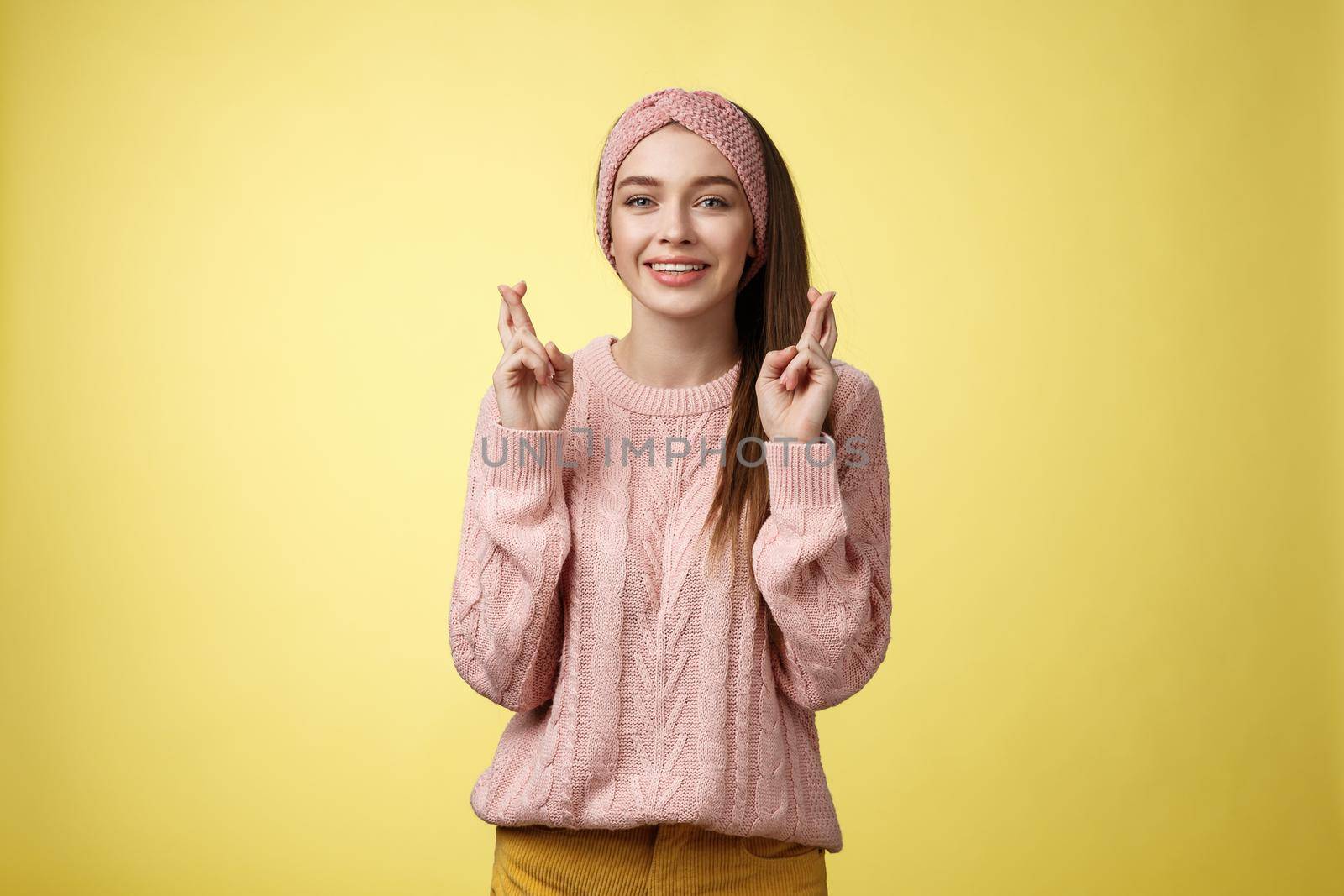 Cross my fingers for you. Supportive cute young european girlfriend wearing sweater, glamour headband making luck, wish gesture smiling hopeful having faith dream come true over yellow background.
