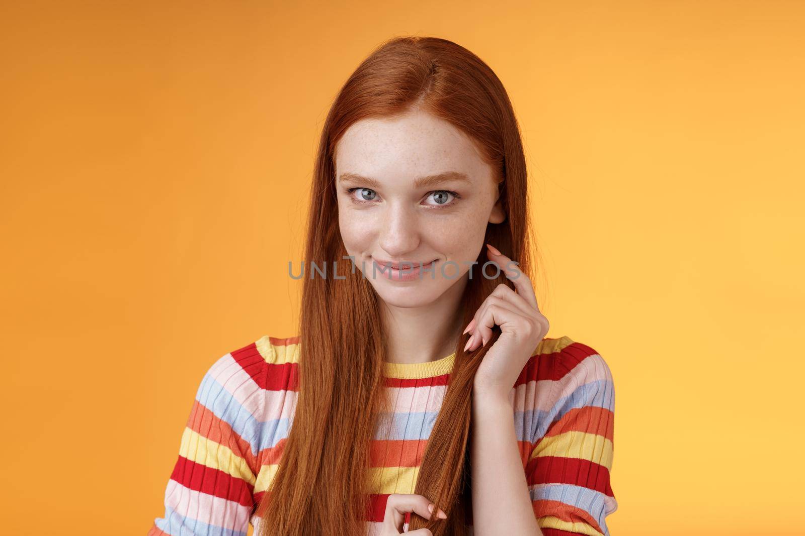 Romantic flirty shy attractive redhead girl 20s touching hair strand smiling silly modest glancing camera coquettish making lovely glances wanna seduce guy expressing sympathy, orange background.