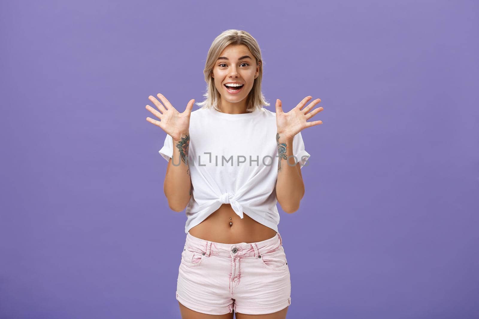 Lifestyle. Indoor shot of delighted and amused attractive stylish urban female with blond hair in stylish white t-shirt and summer shorts raising palms from surprise and amazement standing over purple background.