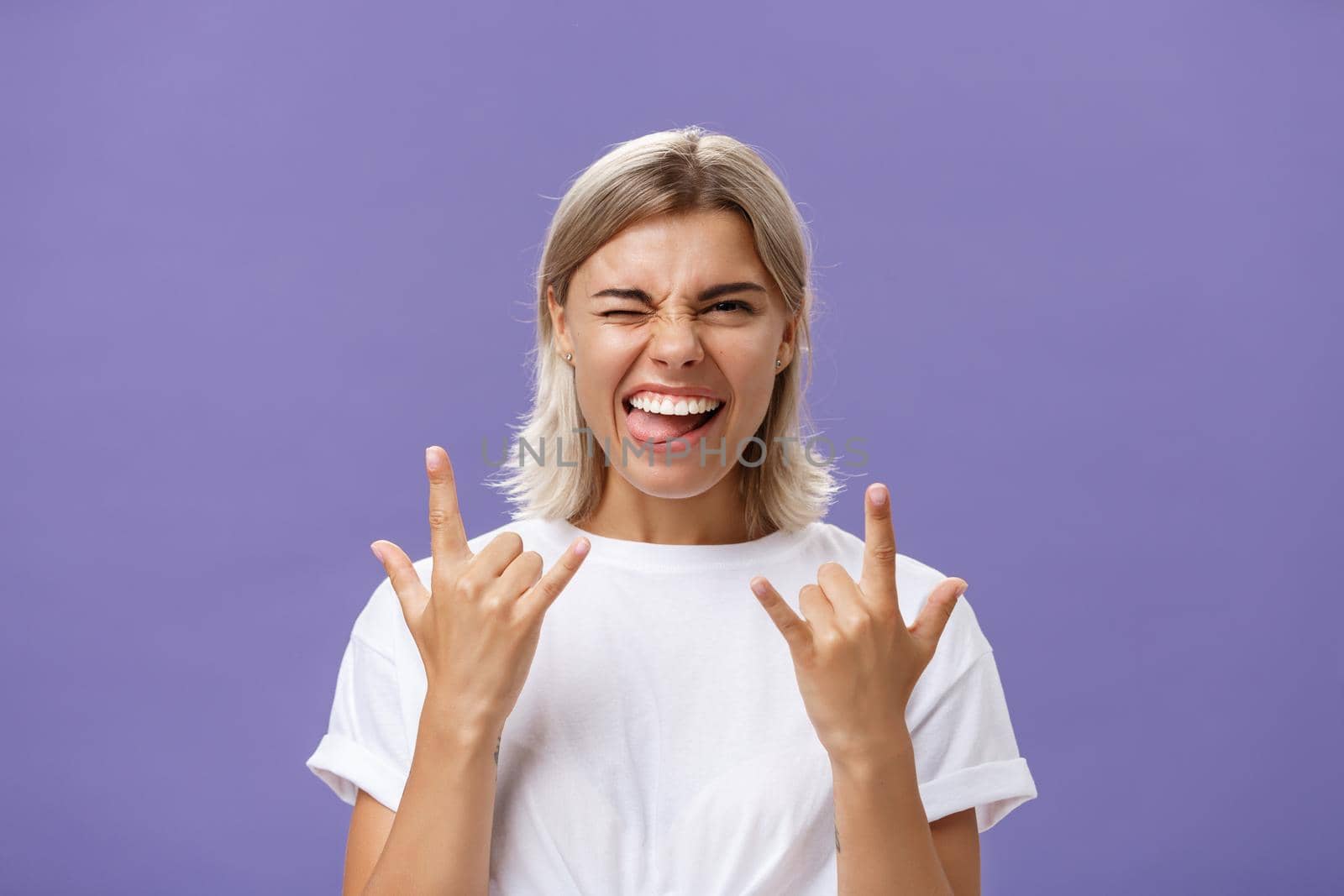 Gonna rock. Portrait of joyful happy good-looking stylish woman with blond medium haircut winking smiling and sticking out tongue while showing rock-n-roll gesture with both hands over purple wall.