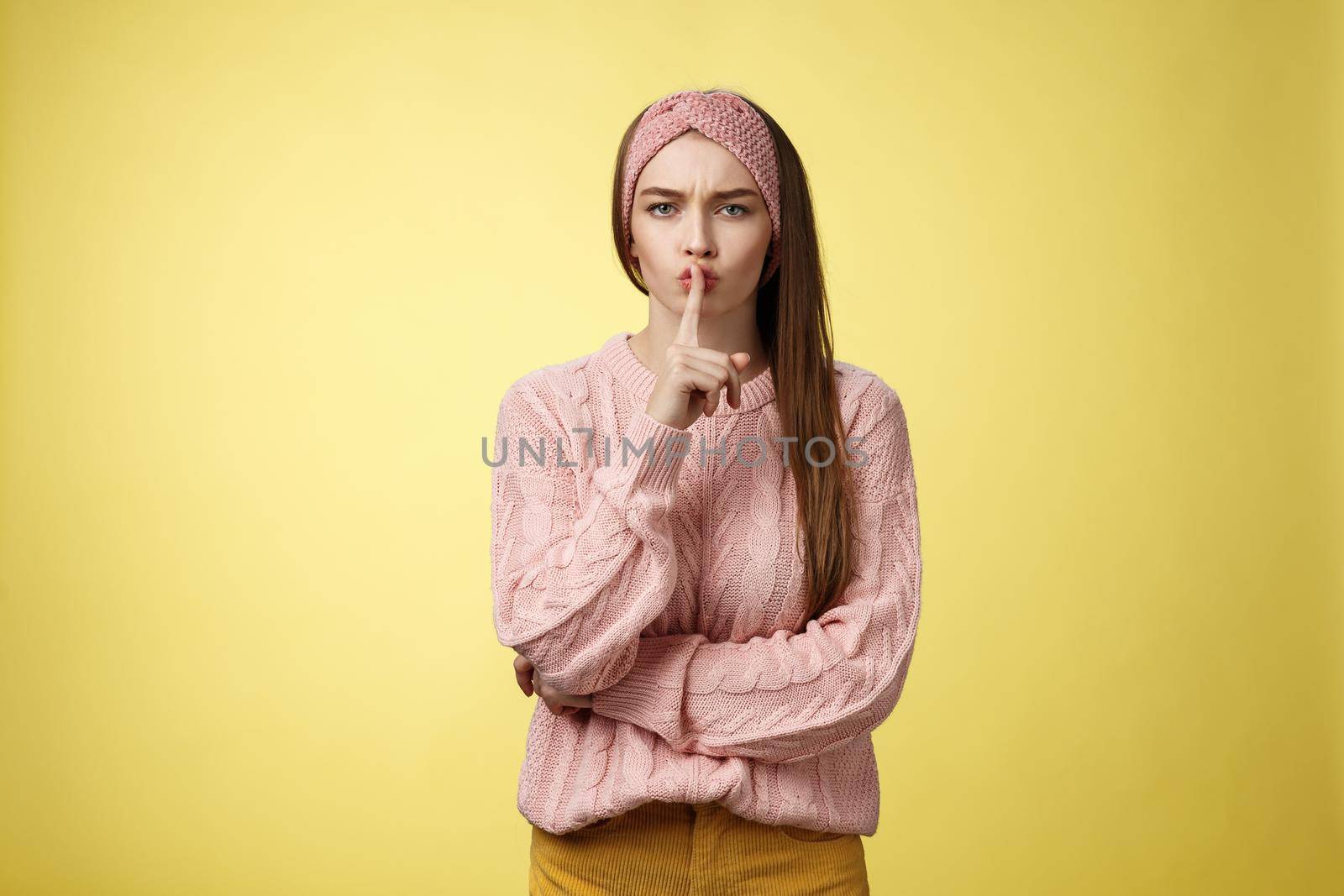Keep mouth shut. Serious-looking bossy attractive young 20s woman in sweater, headband shushing making shhh gesture holding index finger on lips, gossiping, spread rumors over yellow background.