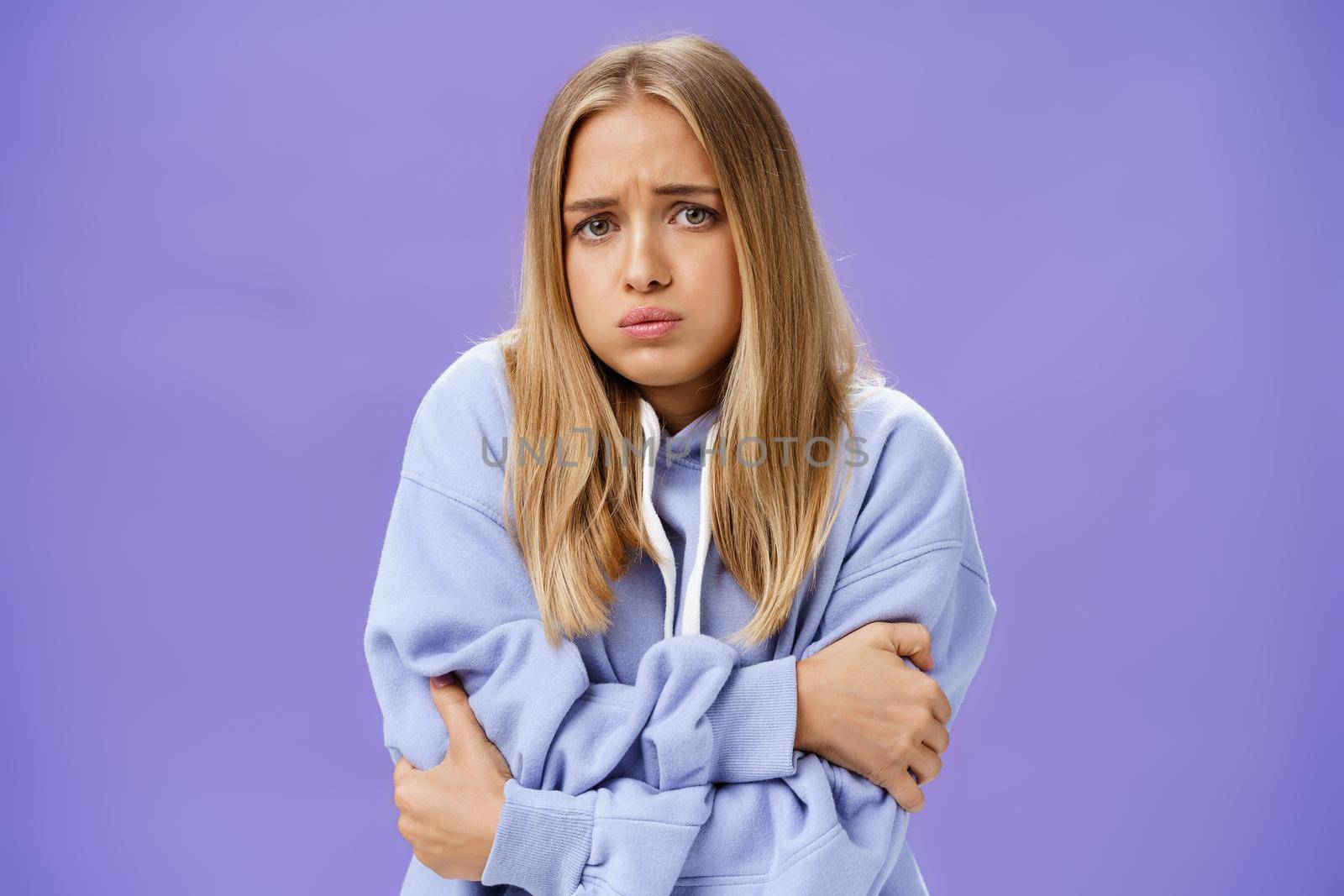 Girl freezing cold during chilly autumn evening trembling hugging herself with hands crossed against chest sulking, frowning wearing hoodie walking on windy and rainy weather posing over purple wall. Clothes, temprature and forecast concept