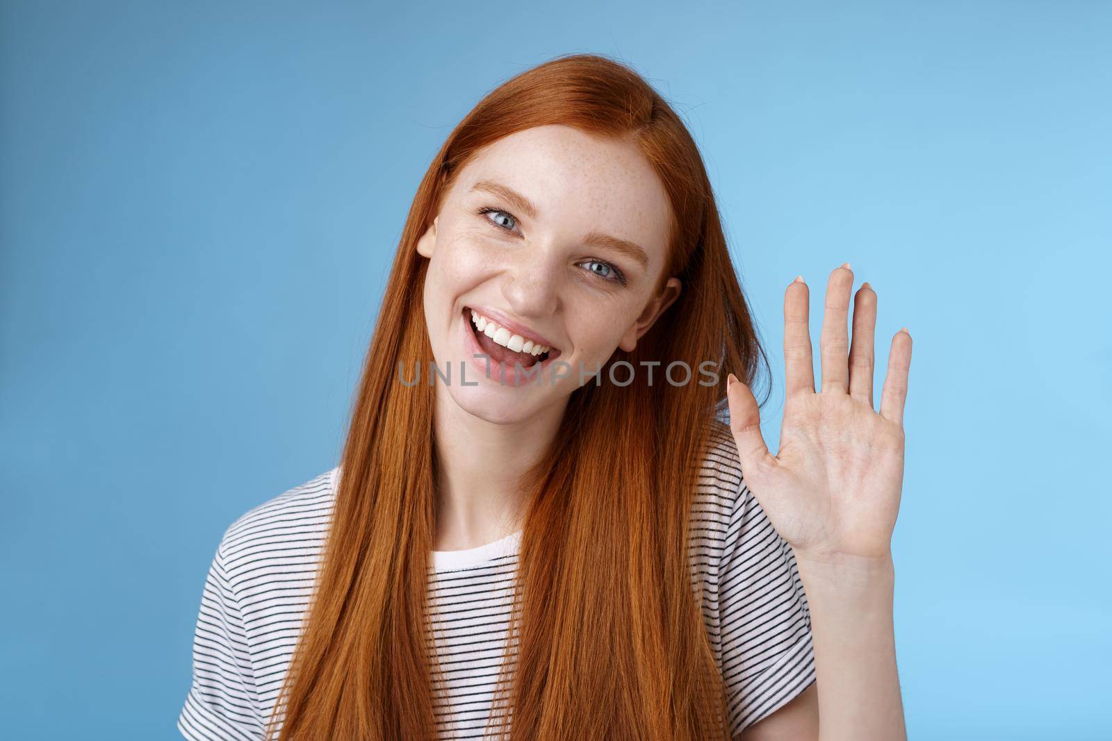 Attractive confident redhead sassy girl pure clean skin blue eyes tilting head cheerfully waving hand hello hi gesture greeting you look camera friendly welcoming friend, standing studio background.