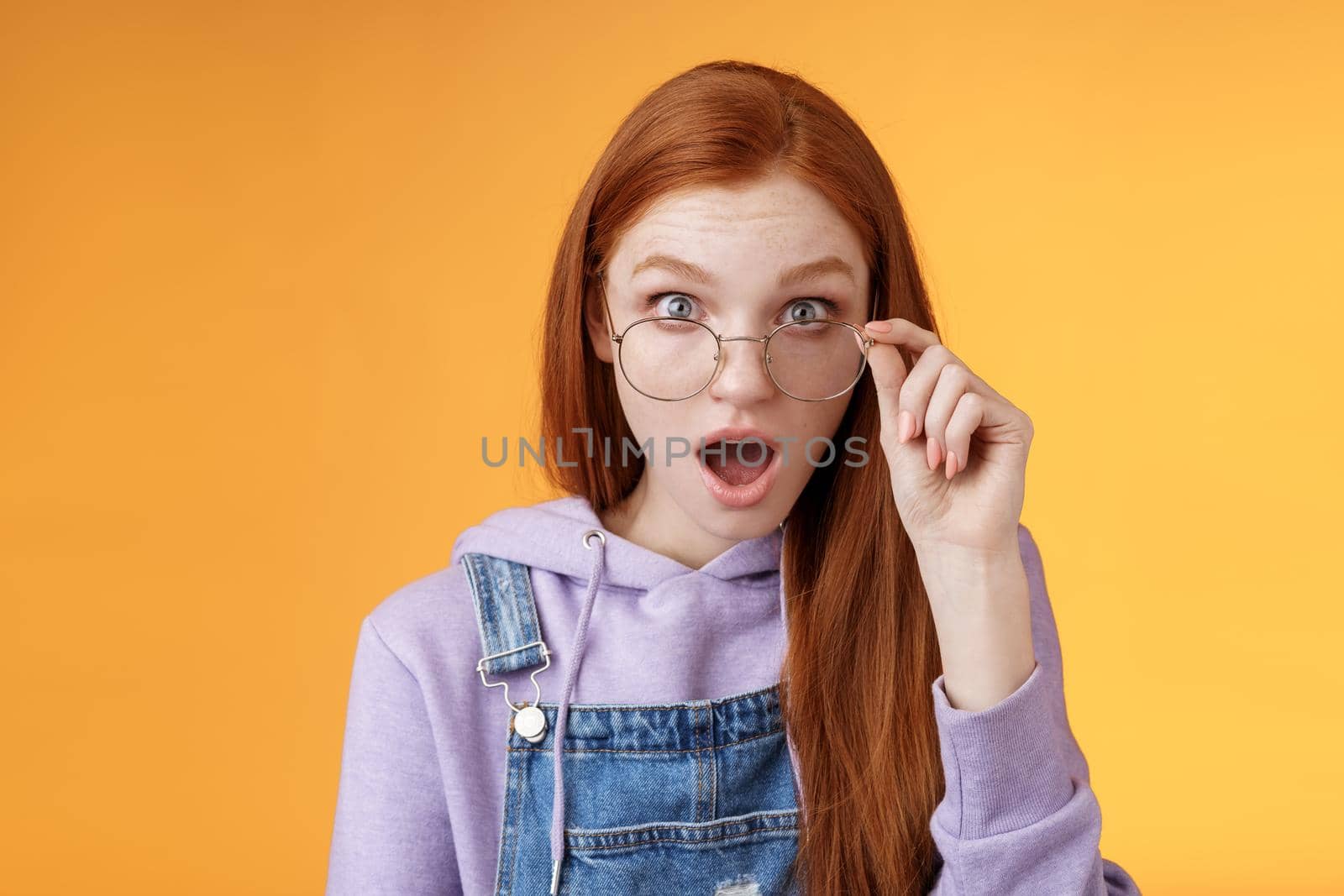 Shocked impressed redhead coworker hearing last intimate rumors drop jaw staring full disbelief camera confused touch glasses seem unsure truth lie standing orange background astonished.