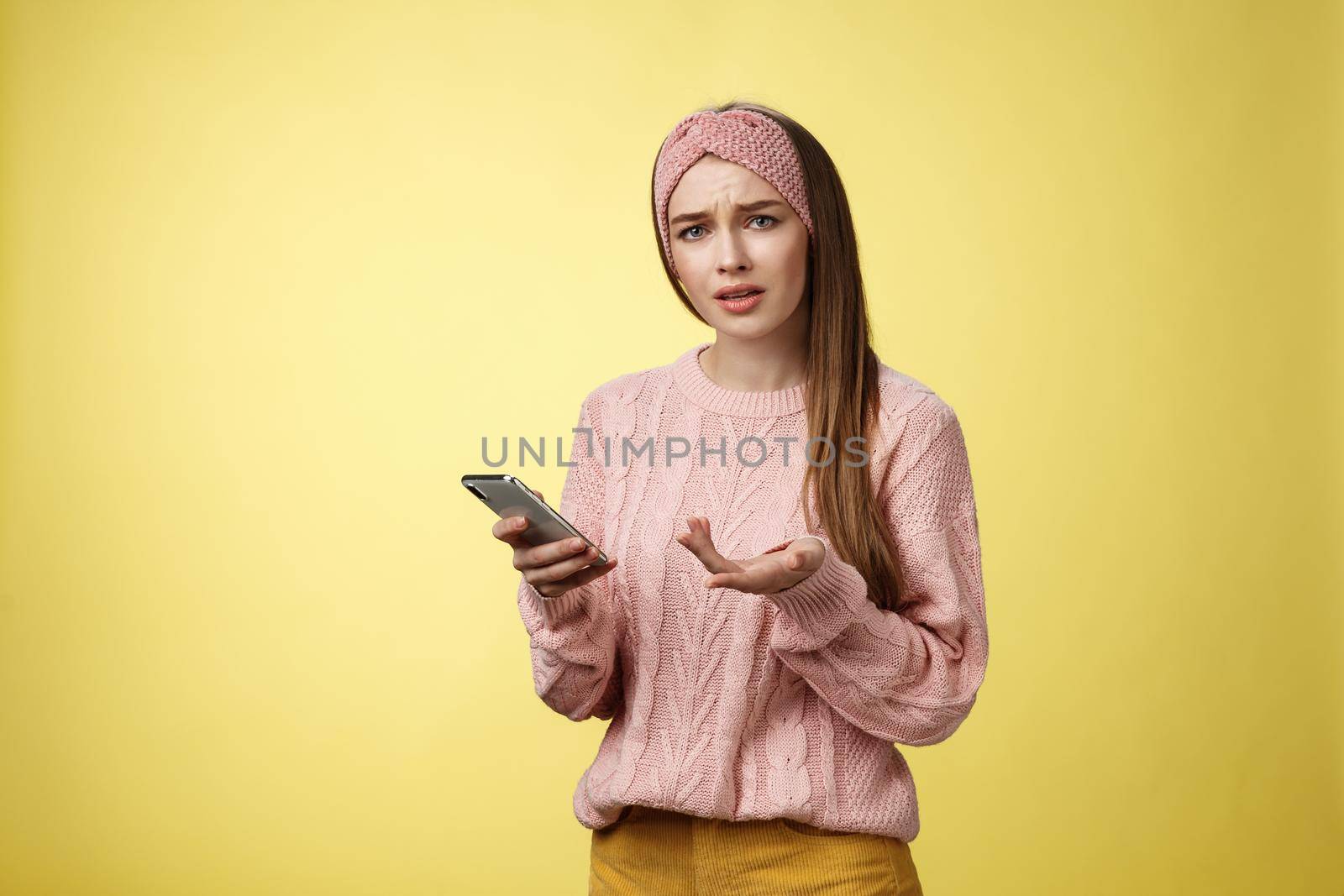 Puzzled confused upset questioned young cute girl in headband knitted sweater frowning clueless raising hand in dismay holding smartphone, cannot understand what happened cellphone over yellow wall by Benzoix