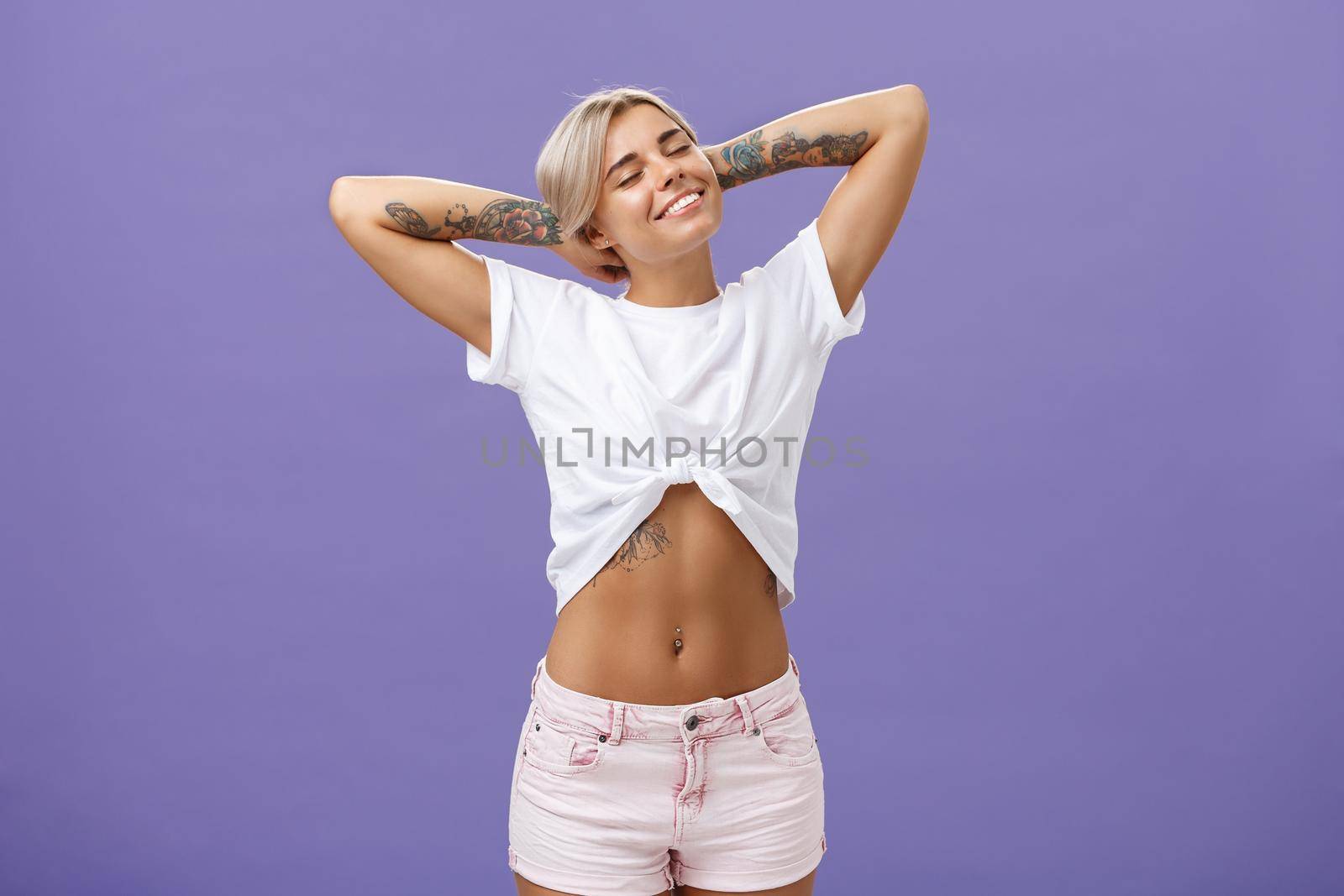 Great feelings after sunbathing. Chill and relaxed attractive young female in cropped top with perfect body and tattoos holding hands behind head, stretching with delight and smiling with closed eyes.