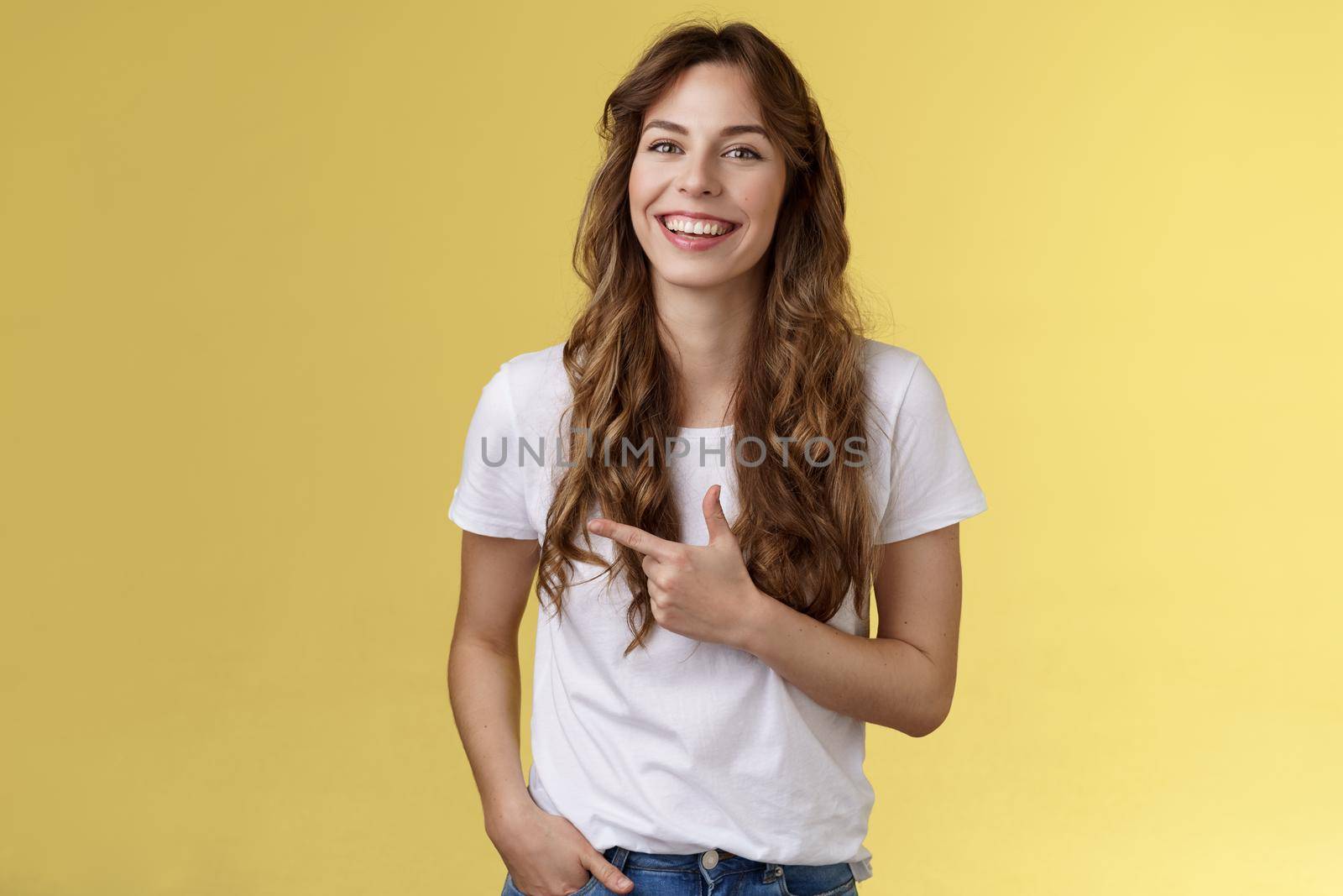 Outgoing charming happy pretty woman curly long hairstyle pointing forefinger left indicate advertisement laughing carefree satisfied hold hand pocket casual friendly pose yellow background.
