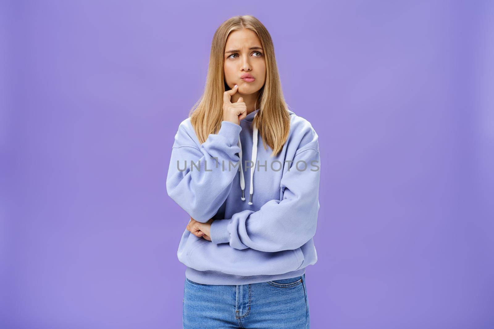 Silly concerned girlfriend trying think up how solve problem standing thoughtful with troubled expression touching jaw pouting, frowning staring right, thinking making up decision over purple wall by Benzoix