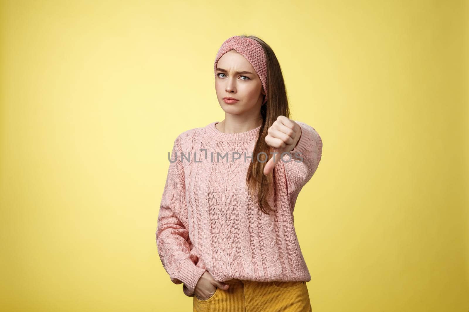 Not cool. Frustrated displeased and disagree cute young woman wearing knitted sweater frowning showing thumb-down smirking judgemental, expressing disapproval of unwell situation over yellow wall by Benzoix