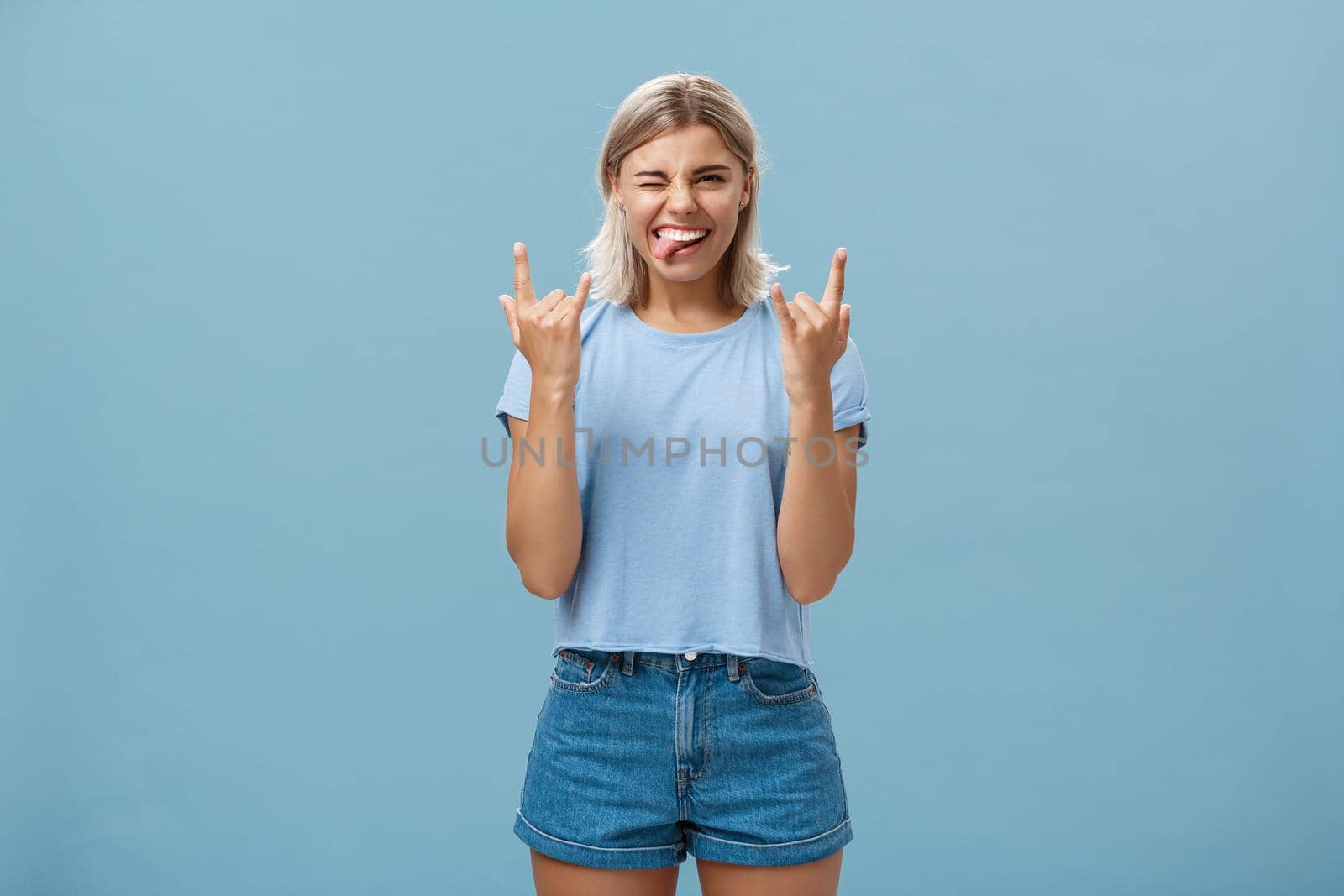 Let us rock this party. Portrait of joyful good-looking and carefree young artistic female musician with blond hair showing heavy metal gesture sticking out tongue and winking amused, having fun.