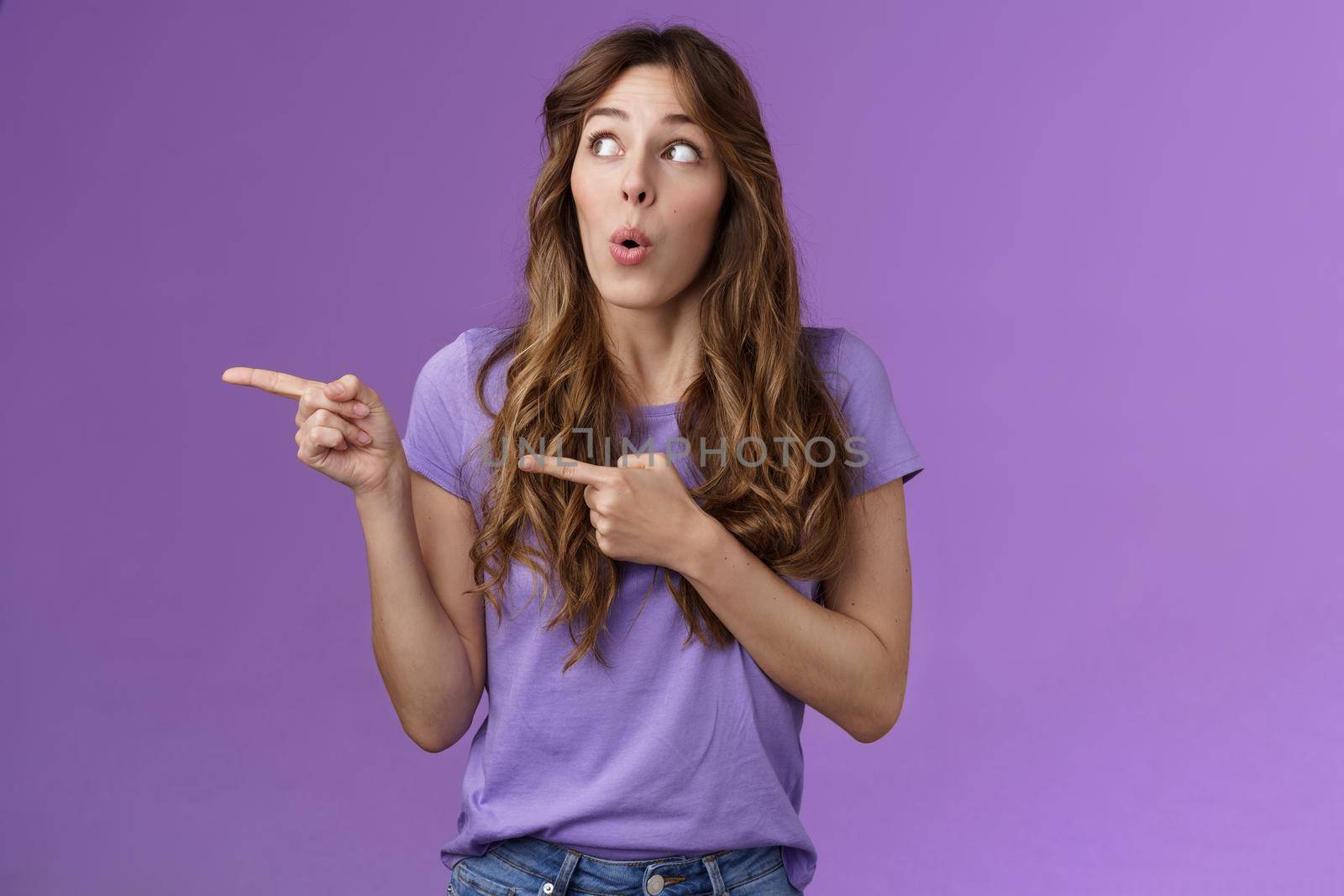 Curious surprised cute curly-haired caucasian female coworker discuss project amazed awesome cool result turn pointing left impressed folding lips wow tempating try new product purple background. Lifestyle.