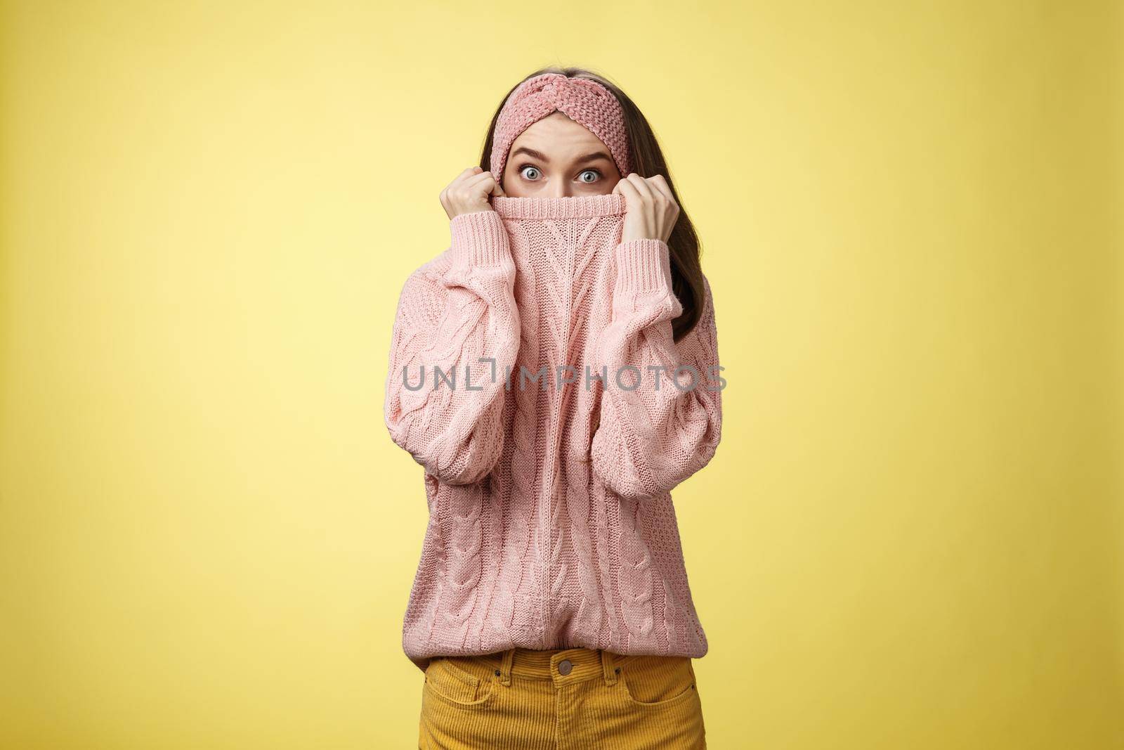 Girl hiding in collar wearing knitted sweater over nose popping eyes, staring bugged eyes at camera, hiding face reacting amazed and shocked, feeling scared, standing in stupor over yellow background.