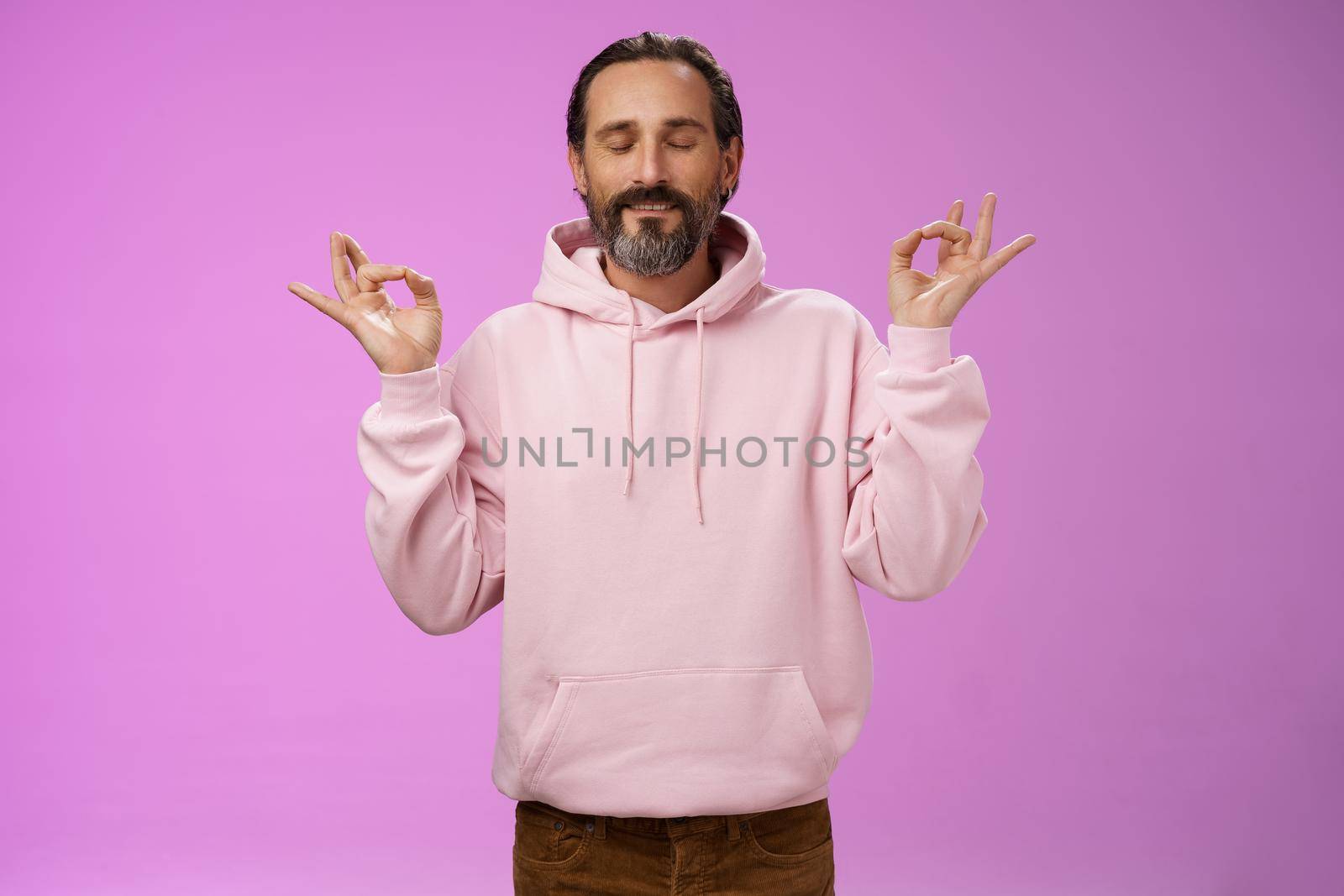 Peaceful charming hipster old man wearing cool pink hoodie close eyes breathing practice release stress meditating standing relaxed happy yoga meditation pose nirvana gesture, purple background.