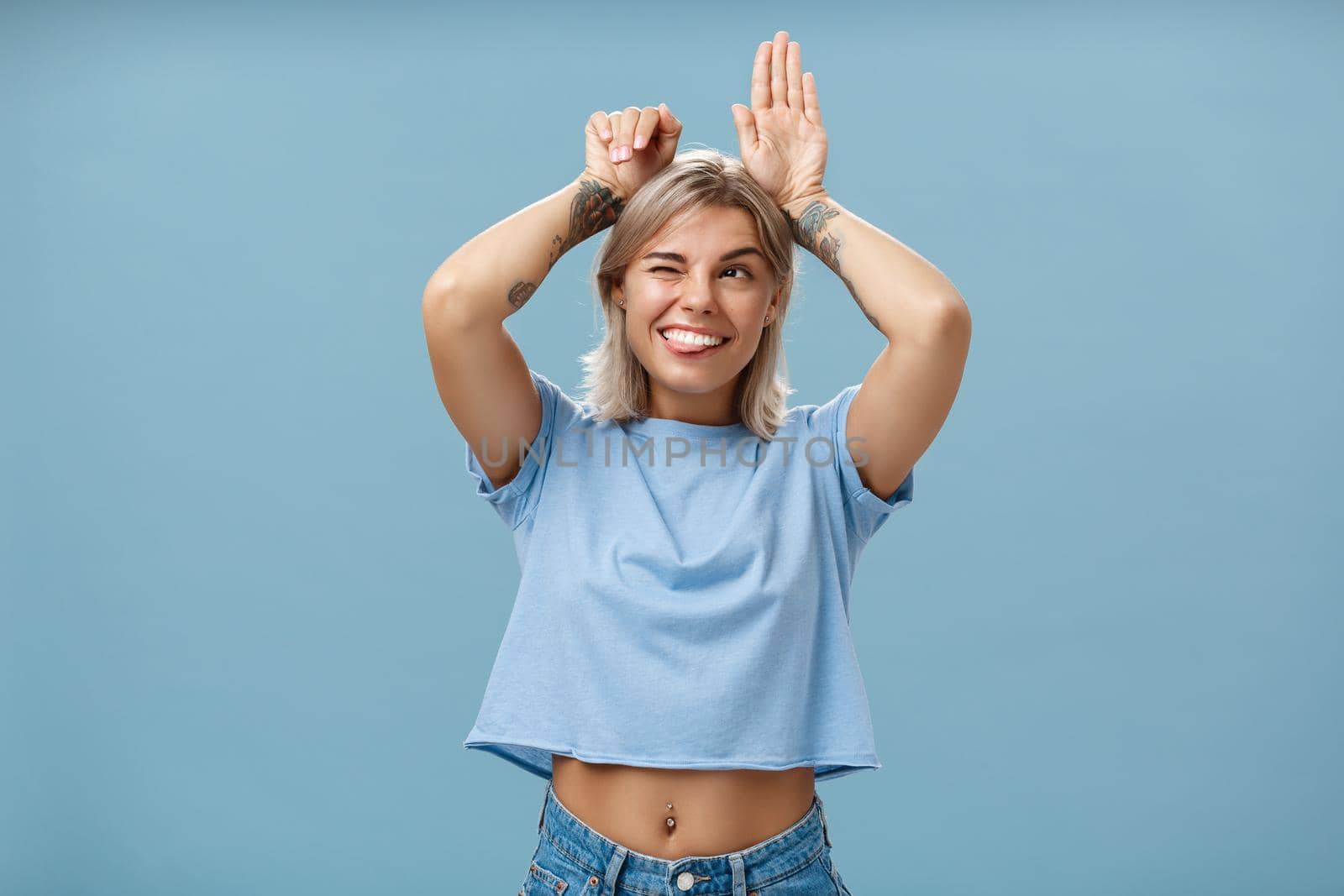 Lifestyle. Studio shot of entertained carefree and emotive happy charming woman with tattoos on arms acting like bunny with palms on head winking joyfully smiling and sticking out tongue over blue wall.