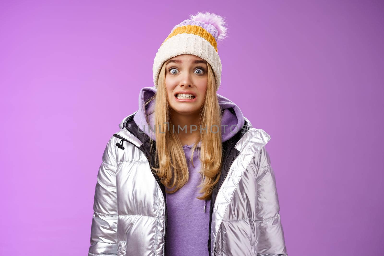 Awkward worried cute timid blond girl in silver jacket hoodie winter hat clench teeth popping eyes camera ooops make mistake standing nervous someone notice, purple background. Copy space