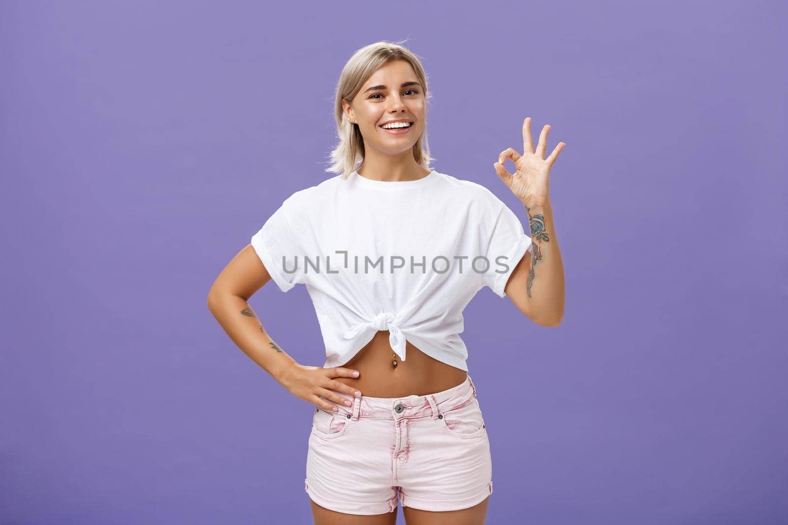Excellent I like it. Portrait of satisfied good-looking happy girl with fair hair in white t-shirt and shorts showing ok or perfect gesture and smiling broadly holding hand on waist over purple wall.