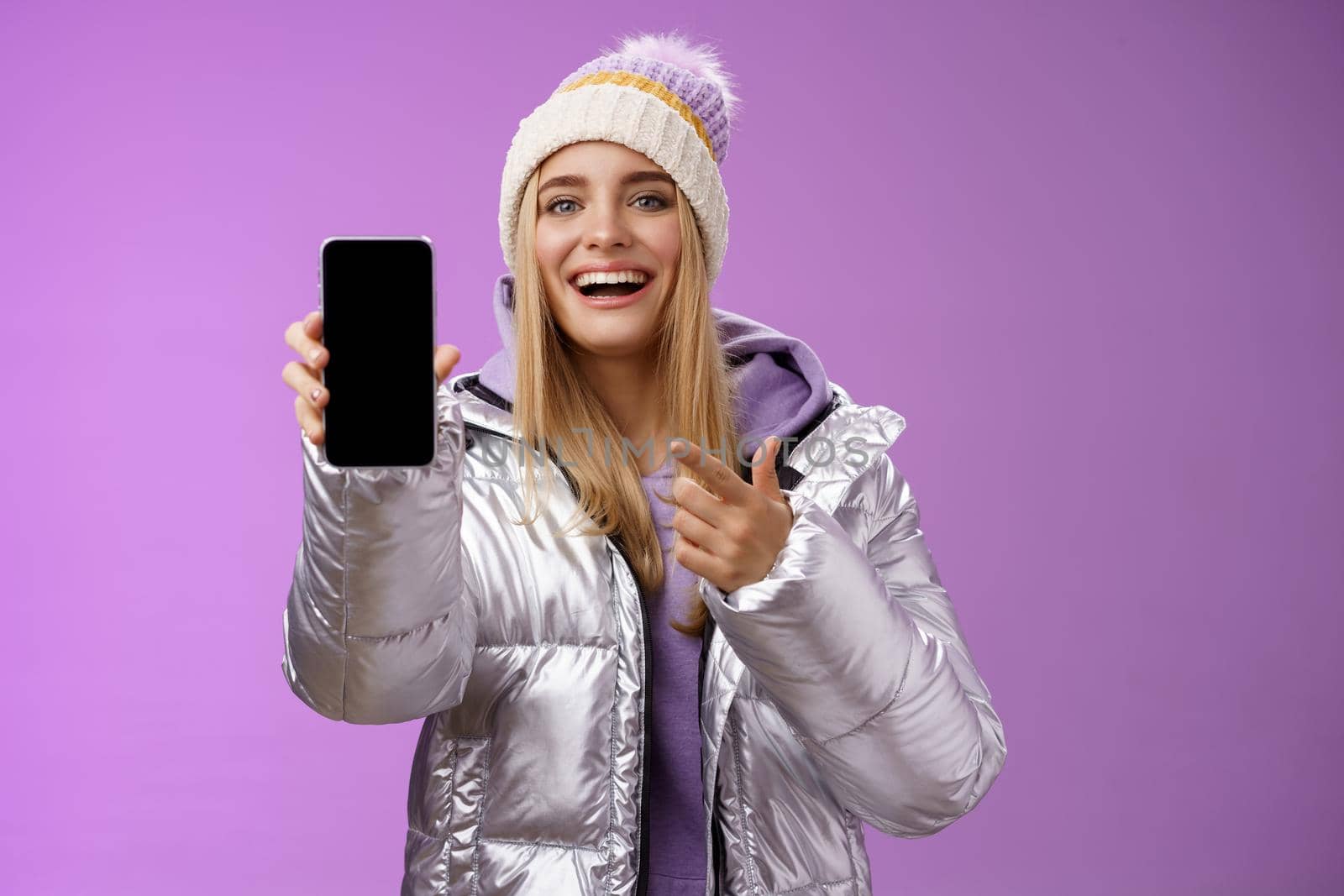 Satisfied amused good-looking blond girl suggest take look smartphone display smiling happily pointing mobile phone delighted talking about awesome new app features, standing purple background by Benzoix