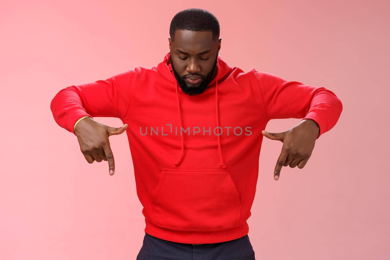 Focused interested serious-looking young african american bearded strong guy wearing red hoodie bend head look pointing down curiously, intrigued what happening lower floor, standing pink background.