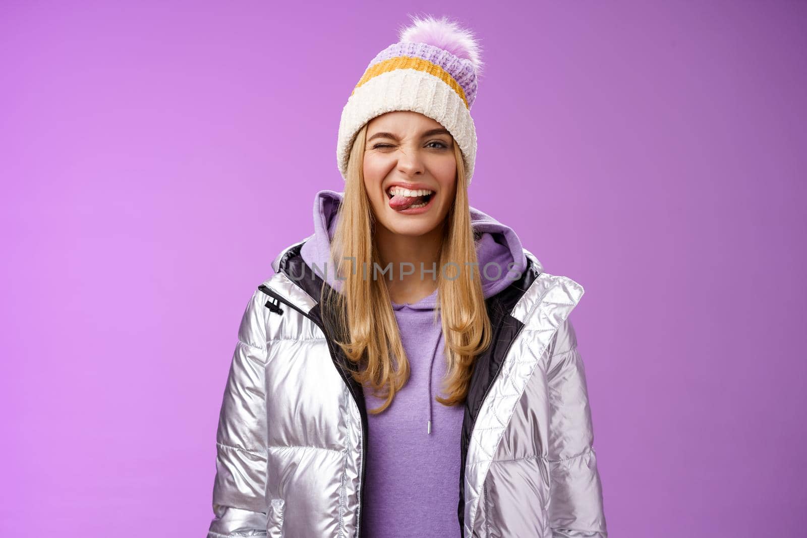 Cheeky carefree delighted charming blond girl having fun feel amused positive show tongue smiling broadly wearing winter silver glittering jacket stylish hat enjoying awesome ski vacation by Benzoix