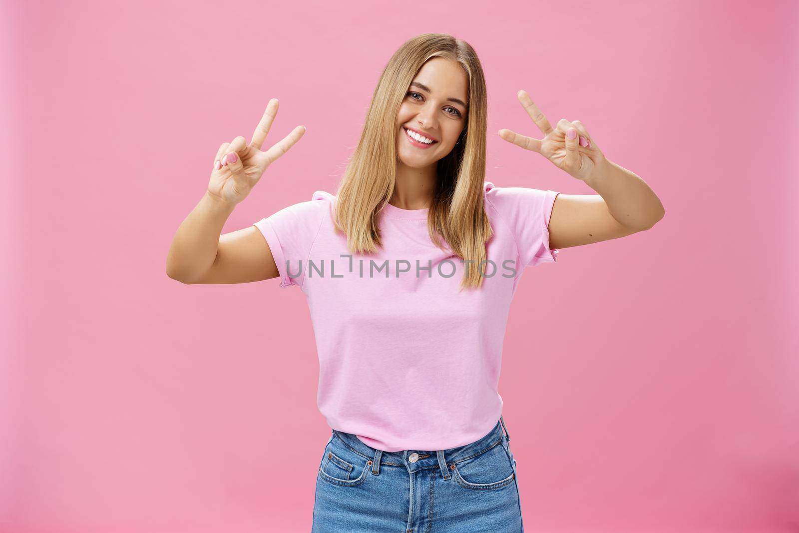 Cute chubby carefree young woman with short fair hair tilting head showing peace gestures with happy friendly smile having fun spending time amused and joyful against pink background. Copy space