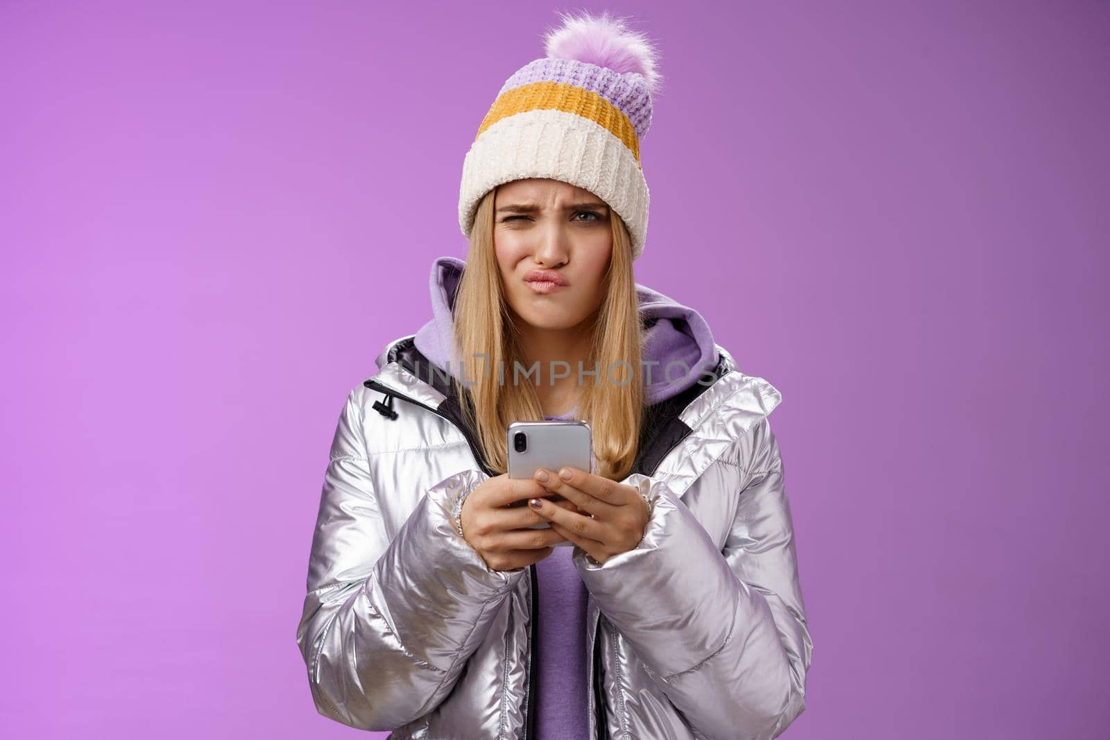Perplexed displeased cute blond stylish girl unwilling anser message cringing reluctant frowning holding smartphone receive disappointed perplexing text, standing unsure purple background.