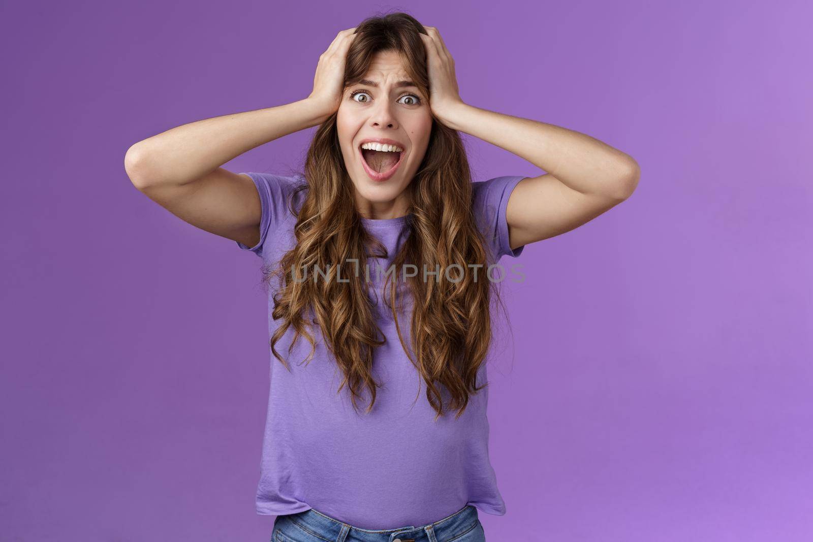 Shocked upset disappointed curly girl shouting sorrow face troublesome shocking situation grab head scream panic outraged pissed and angry, standing distressed purple background. Copy space