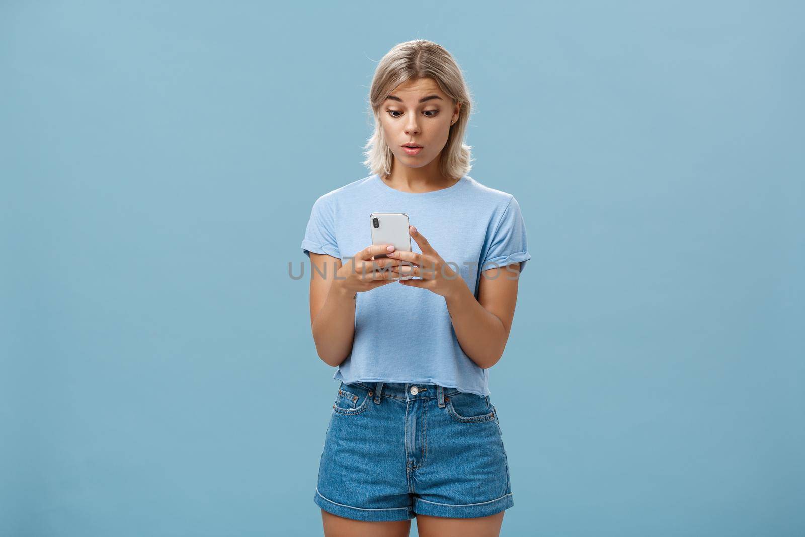 Lifestyle. Indoor shot of surprised young caucasian girl receiving unexpected invitation via smartphone reading strange message in cellphone staring stunned and confused at screen over blue background.