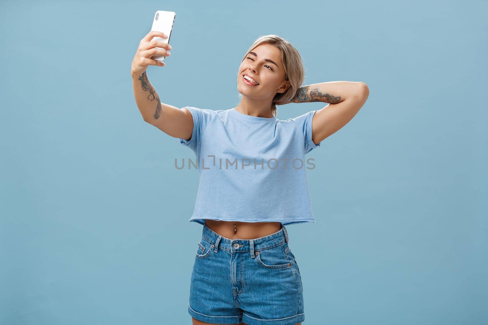 New post in my blog. Stylish feminine and sociable good-looking young female posing for selfie with pulled arm and smartphone sticking out tongue while making faces at device screen over blue wall by Benzoix