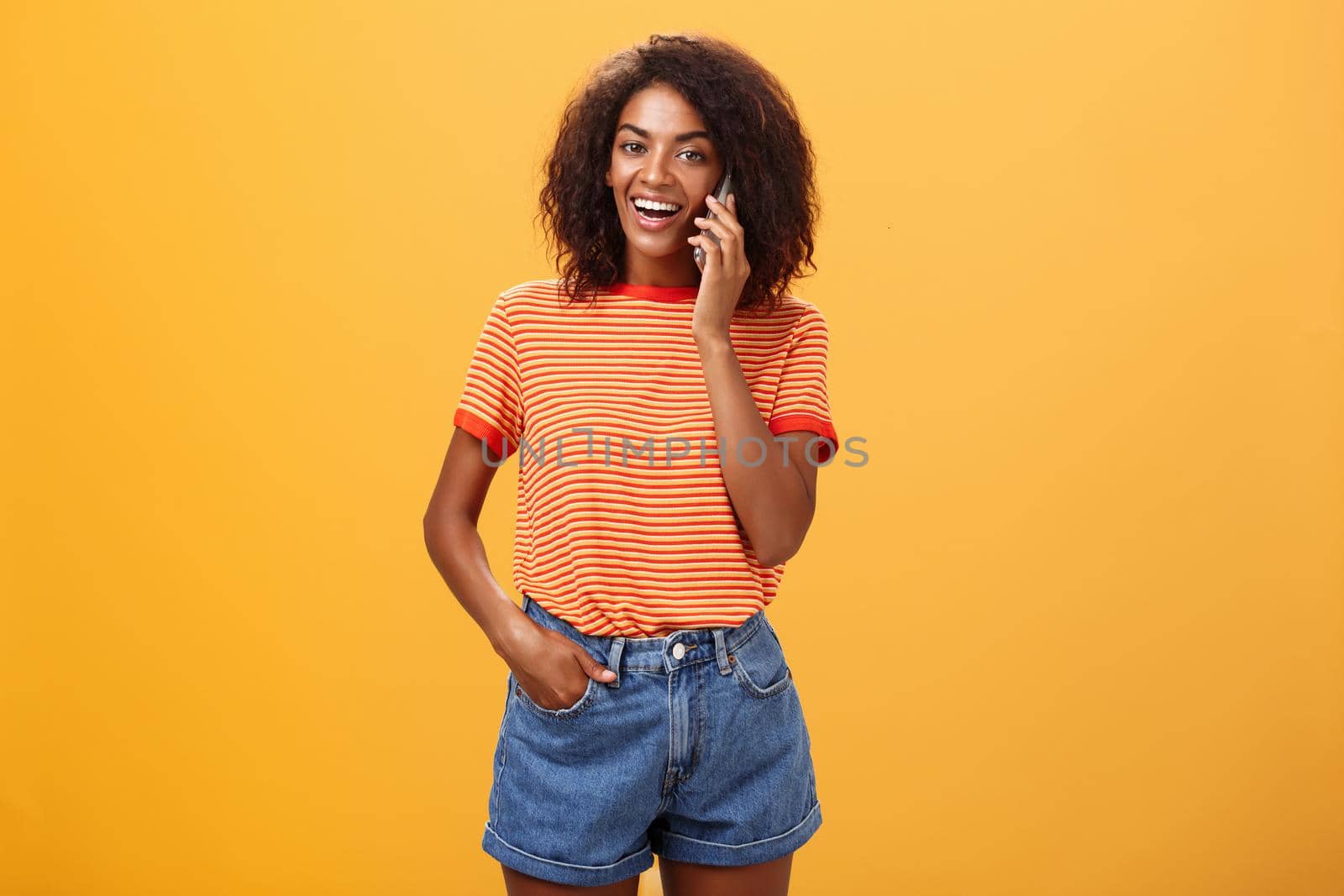 Indoor shot of stylish confident female coworker in trendy t-shirt and denim shorts holding smartphone near ear calling friend talking via cellphone casually about girls stuff over orange background. Lifestyle.