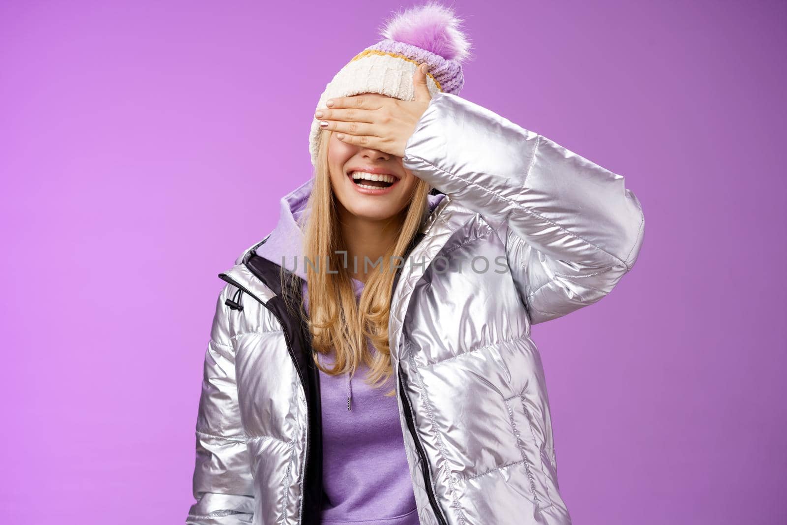 Happy relaxed smiling attractive young blond woman in stylish warm silver jacket hat hide eyes behind palm laughing happily waiting boyfriend prepared surprise gift amused, standing purple background.