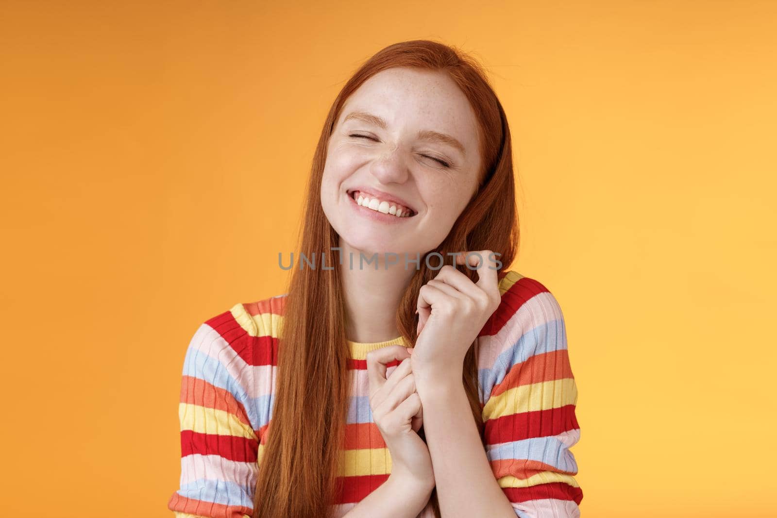 Happy dreamy romantic young tender ginger girl fantasizing creating love story imagination smiling broadly delighted close eyes touching hair strands recalling nice memory, standing orange background by Benzoix