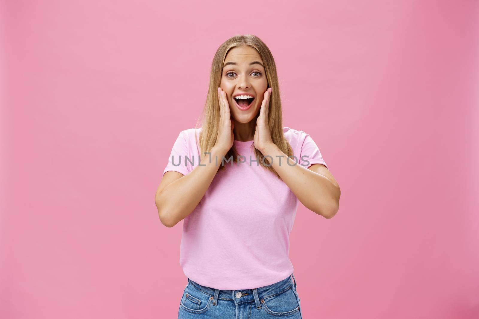Delighted and amazed happy girlfriend in t-shirt and jeans smiling broadly with opened mouth amused touching cheeks surprised and satisfied reacting to positive surprise over pink background. Copy space