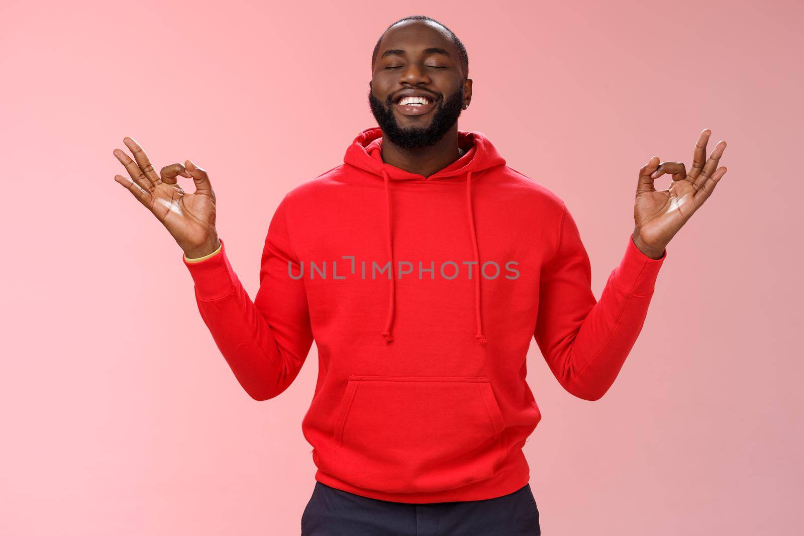 Happy african american bearded man in red hoodie meditating found peace nirvana smiling delighted close eyes relaxed relieved standing lotus mudra pose search zen, practice yoga, pink background by Benzoix