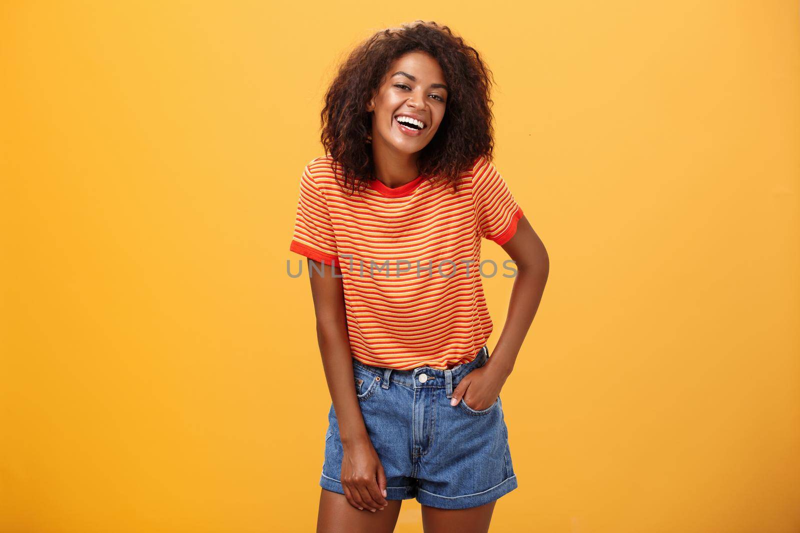 Portrait of charismatic charming african american stylish african american female in trendy shorts and t-shirt laughing happily enjoying talking with cool people laughing posing over orange background. Lifestyle.