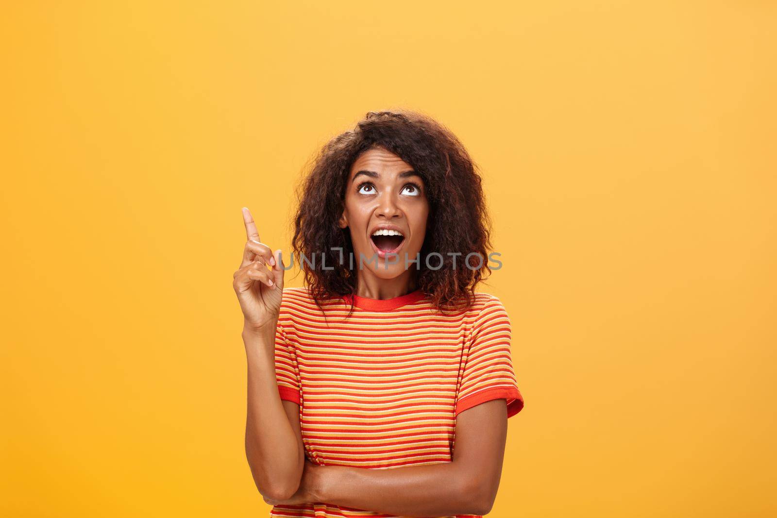 Waist-up shot of interested curious good-looking dark-skinned female in striped t-shirt talking asking question about curious star looking and pointing up with joy over orange background by Benzoix