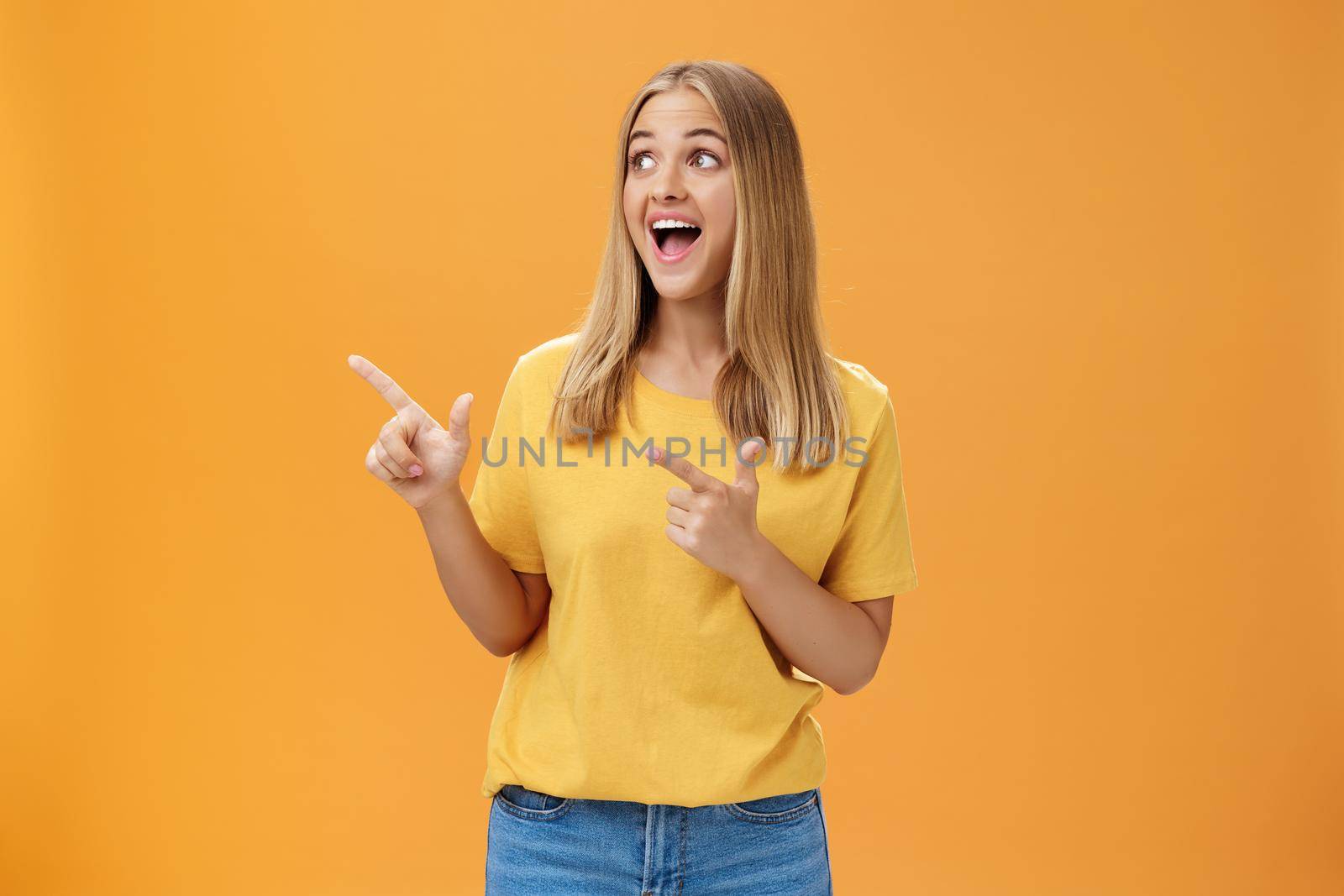 Amused enthusiastic and charismatic girl with blond hair and tanned pure skin without makeup pointing and looking at upper left corner with joyful happy smile over orange background by Benzoix