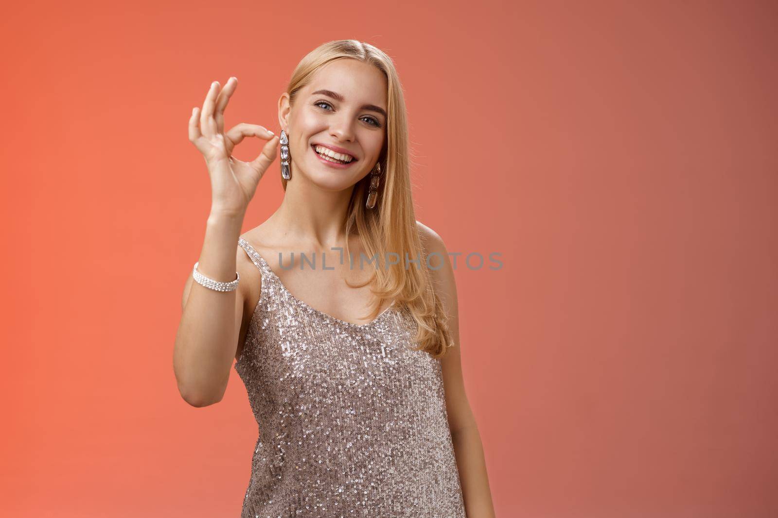 Optimistic attractive stylish blond woman in silver dress show okay ok gesture smiling broadly give approval like recommend cool store buy outfit birthday party, standing red background peleased.