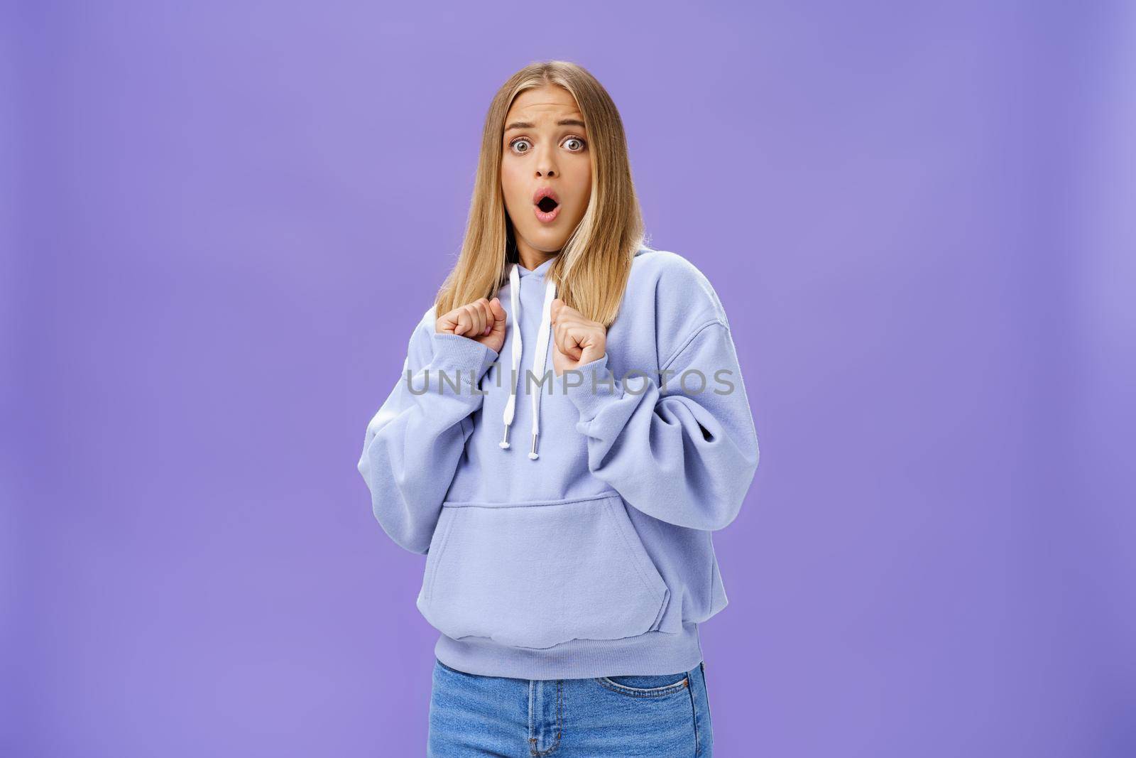 Woman scared of sudden loud noise in dark gasping looking shocked and concerned tilting backwards with frightened look pressing clenched fists to body from fear, wearing warm hoodie over purple wall by Benzoix