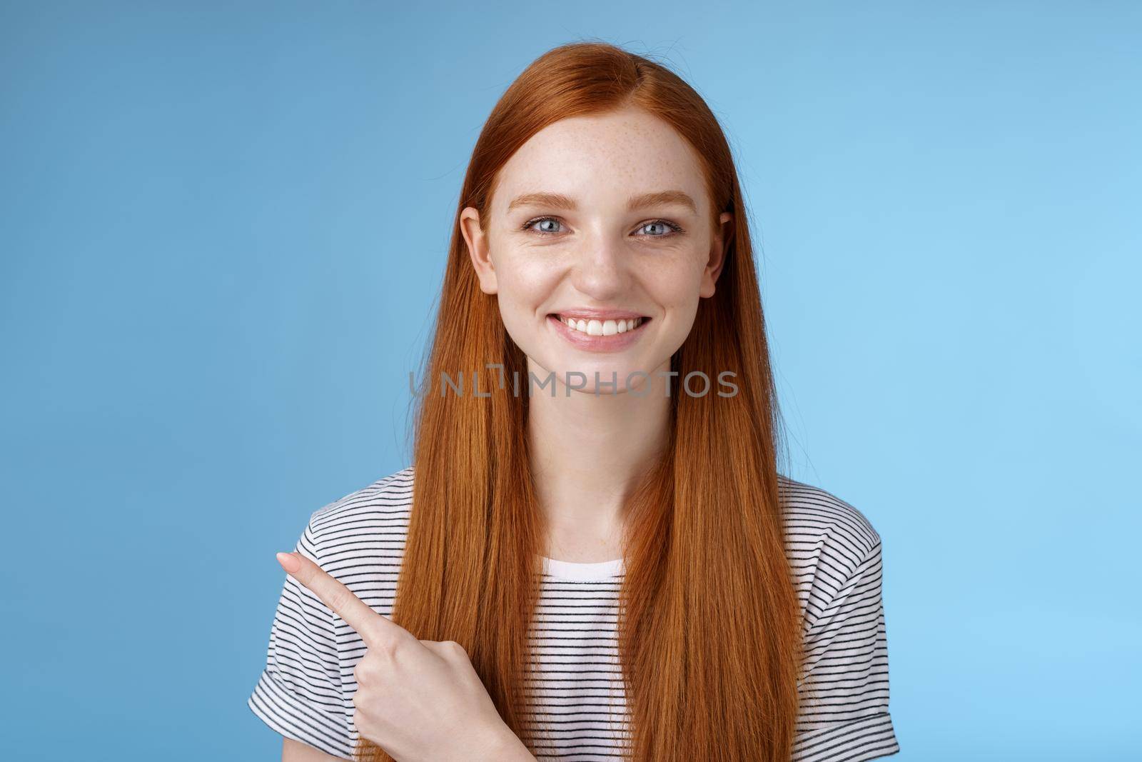 Pleasant charismatic helpful friendly-looking female redhead blue eyes pointing left index finger help make decision pick product store smiling broadly confident gaze camera asking trust her by Benzoix