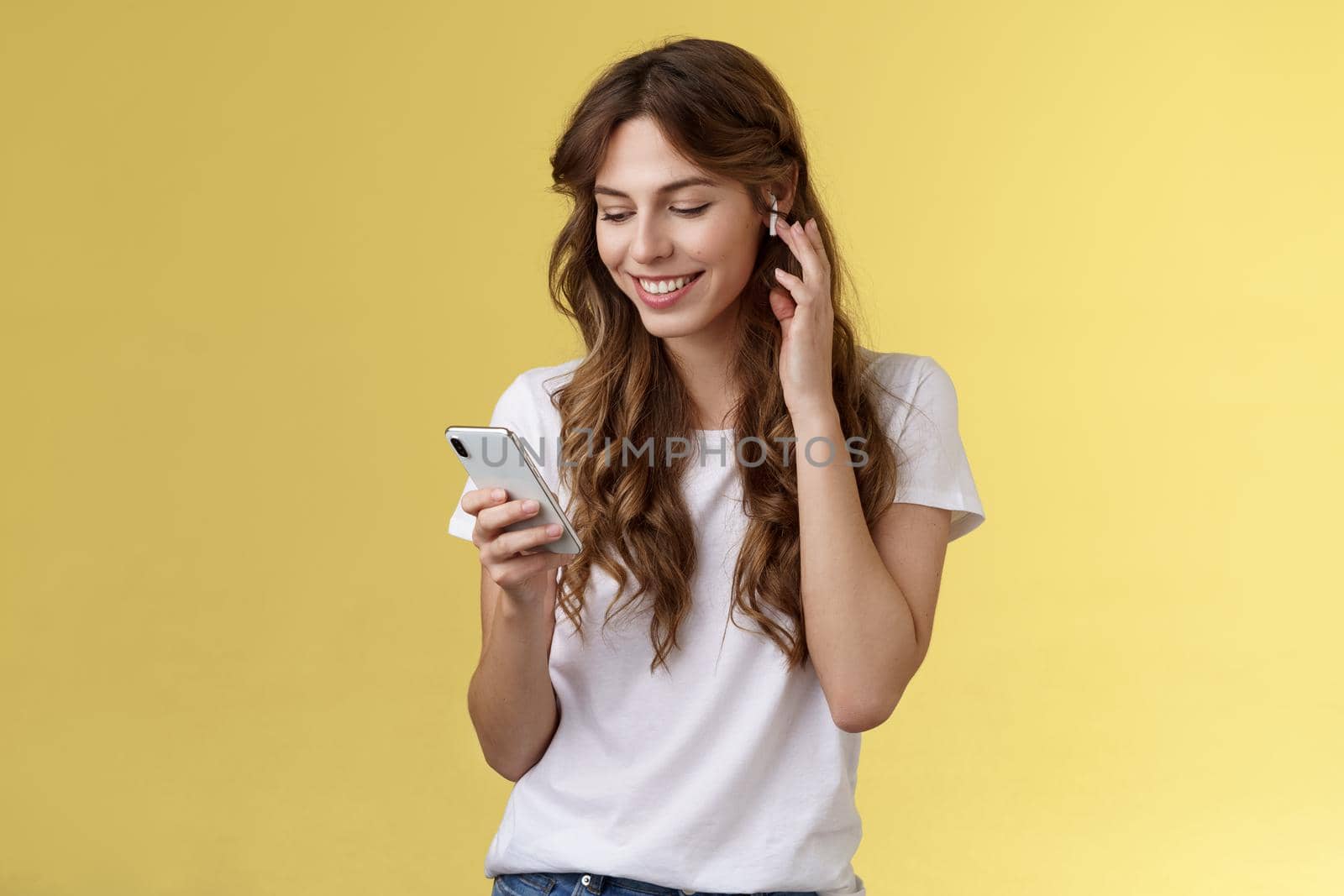 Tender feminine curly-haired female white t-shirt touch wireless earbud put earphone ear smiling pleased look smartphone screen pick song wanna listen music search right track yellow background by Benzoix