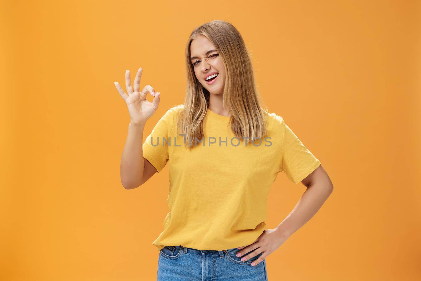 Confident and skillful female student assuring teacher she do great job holding hand on waist winking at camera with self-assured look and showing okay gesture assuring everything ok over orange wall by Benzoix