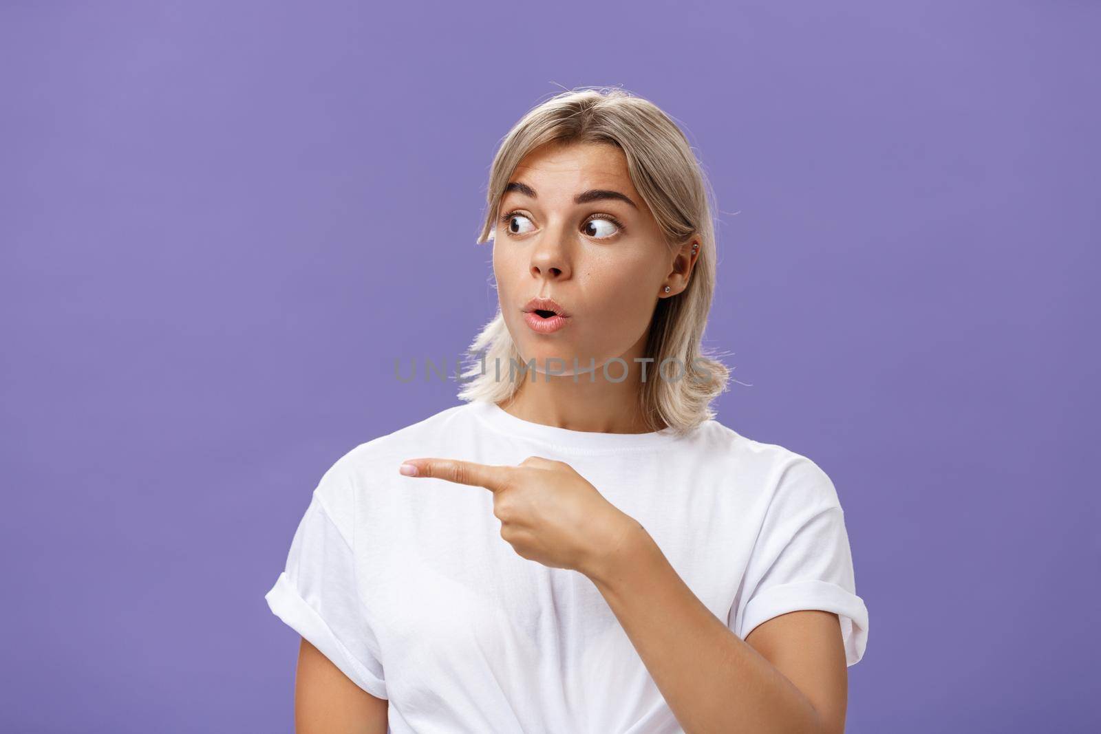 Close-up shot of intrigued interested attractive adult blonde female in white t-shirt folding lips staring and pointing left with curious and thrilled expression over purple background. Advertisement concept