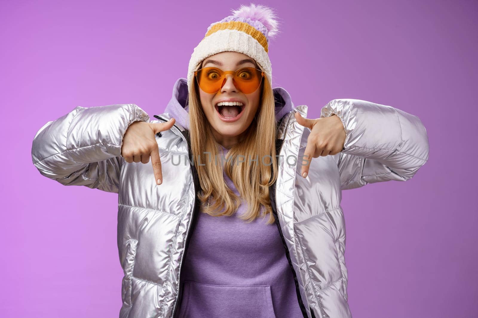 Excited impressed good-looking blond girl drop jaw amused overwhelmed pointing down index fingers checking out awesome promotion standing surprised thrilled wearing silver winter jacket hat.