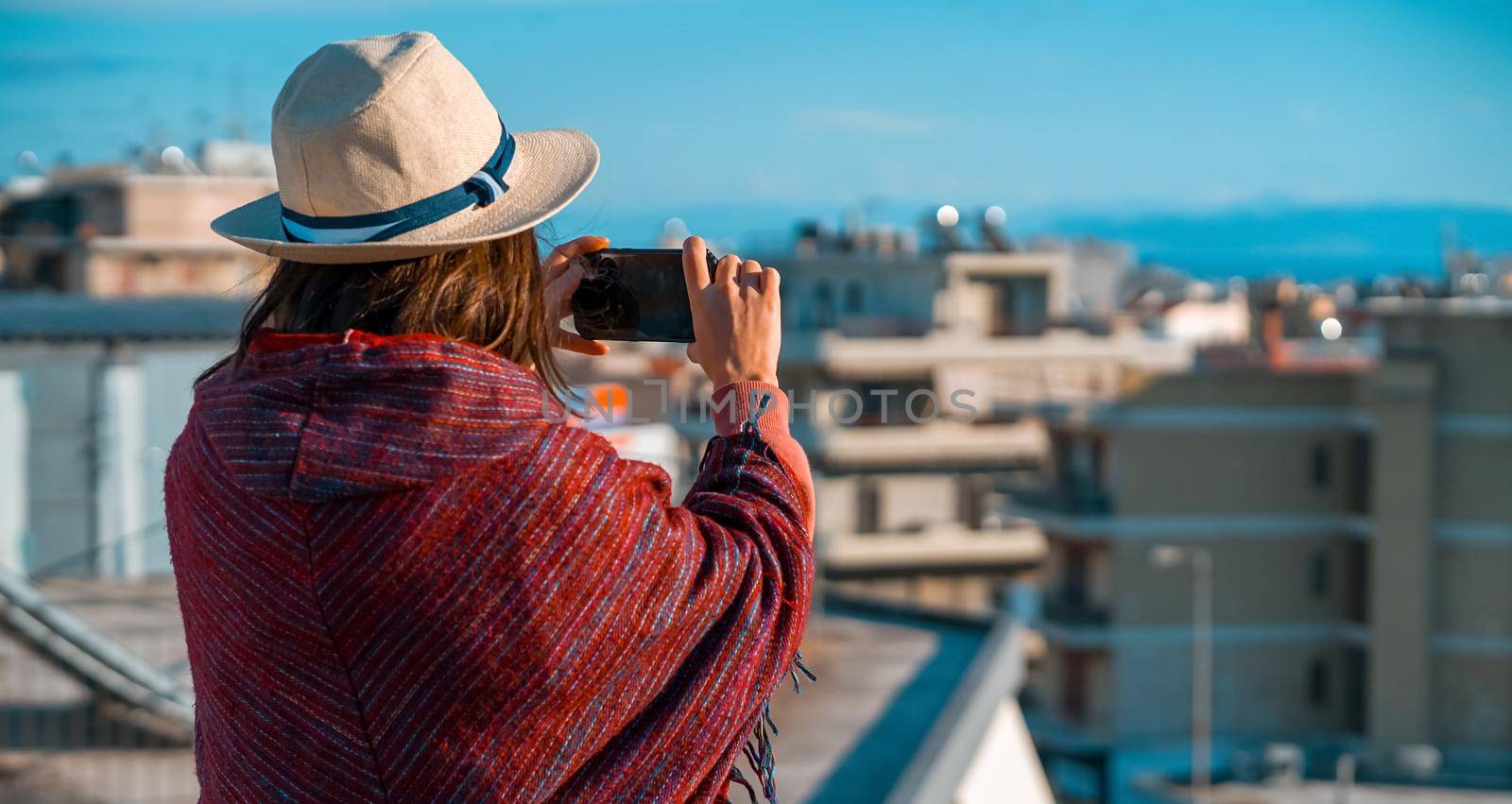 A young girl in a burgundy poncho and a hat travels and takes a photo with a cityscape on a sunny day.