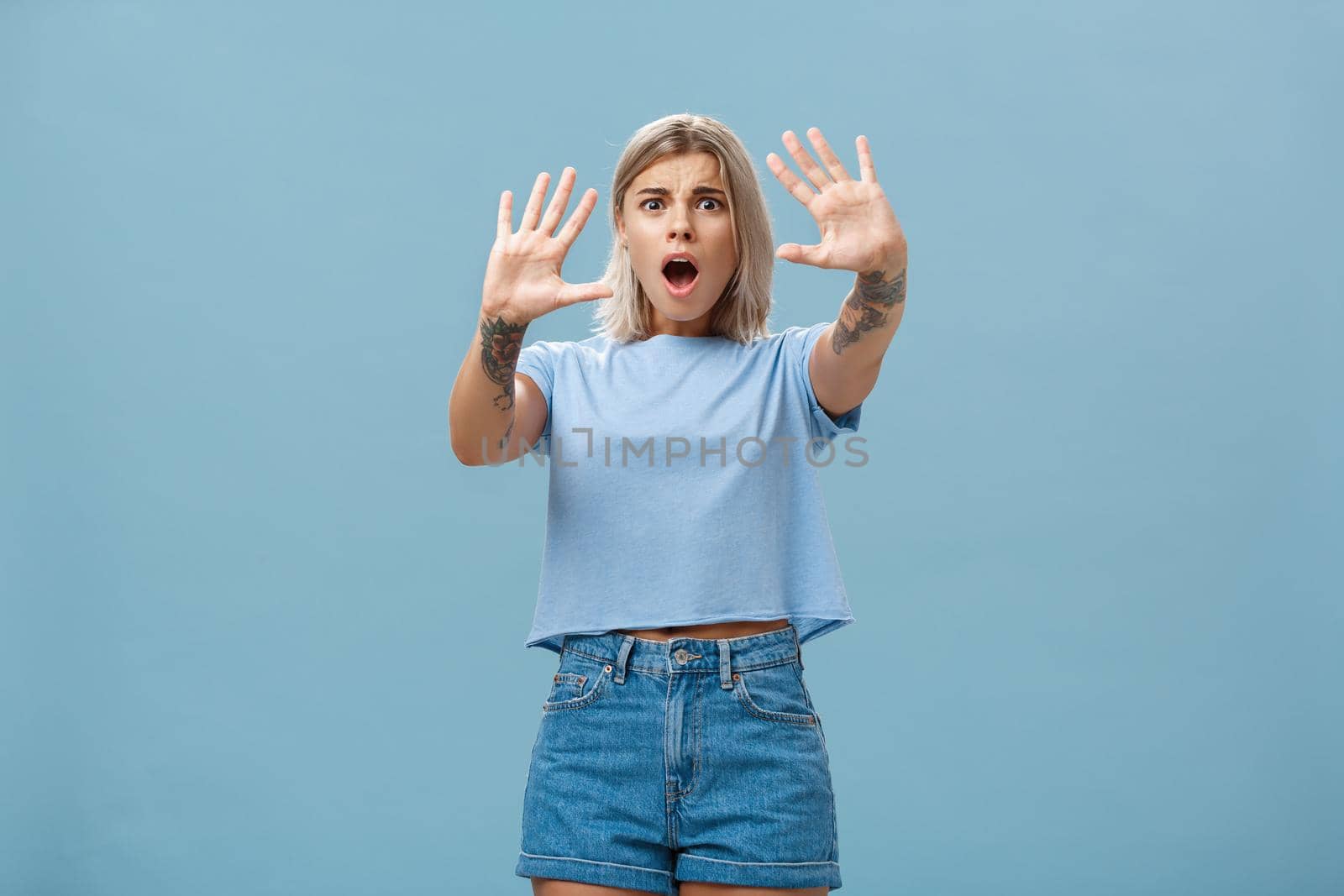 Woman terrified screaming and asking stop. Portrait of shocked panicking troubled blonde female in denim shorts and casual t-shirt pulling hands in no gesture dropping jaw and frowning by Benzoix