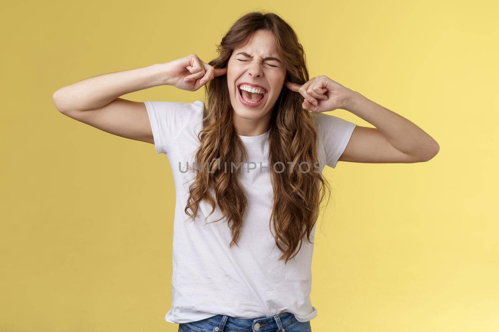 Girl yelling annoyed demand stop playing guitar. Bothered irritated woman close eyes screaming disappointed plug index fingers ear holes disgusted loud terrible noise music stand yellow background.