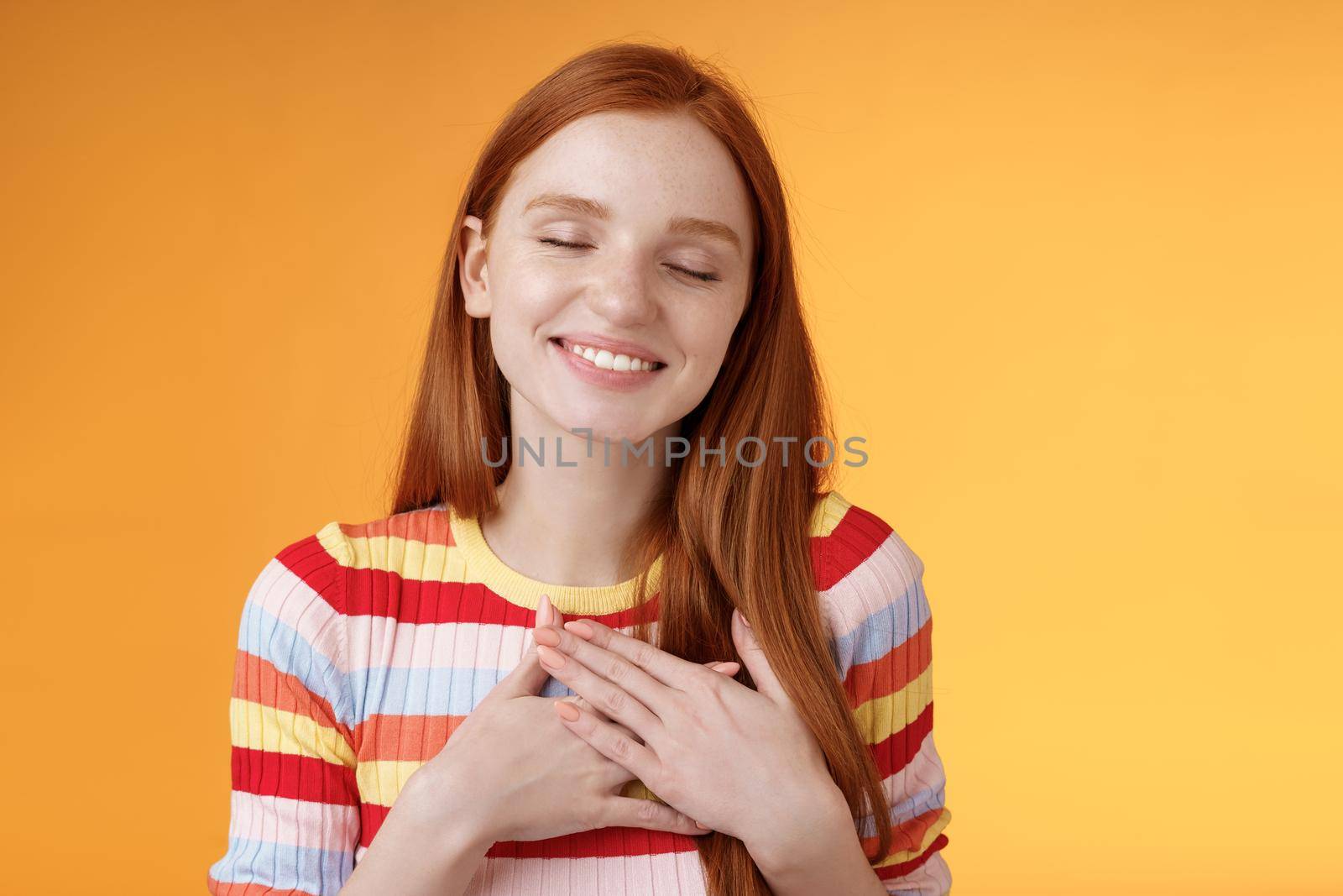 Happy romantic delighted sighing young lovely redhead girlfriend smiling broadly touching heart chest palms close eyes recalling date passionate kiss feel gratitude warmth, orange background.