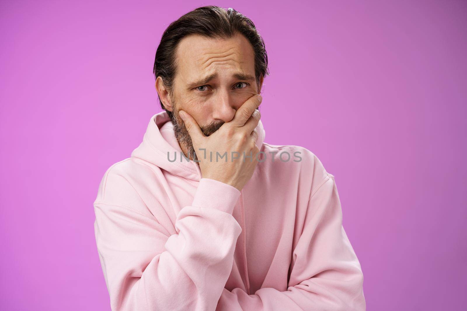 Shocked upset handsome mature bearded man hear terrible news griefing suffer sorrow express empathy gasping hold hand mouth reacting lose cringing crying cannot control emotions, purple background.