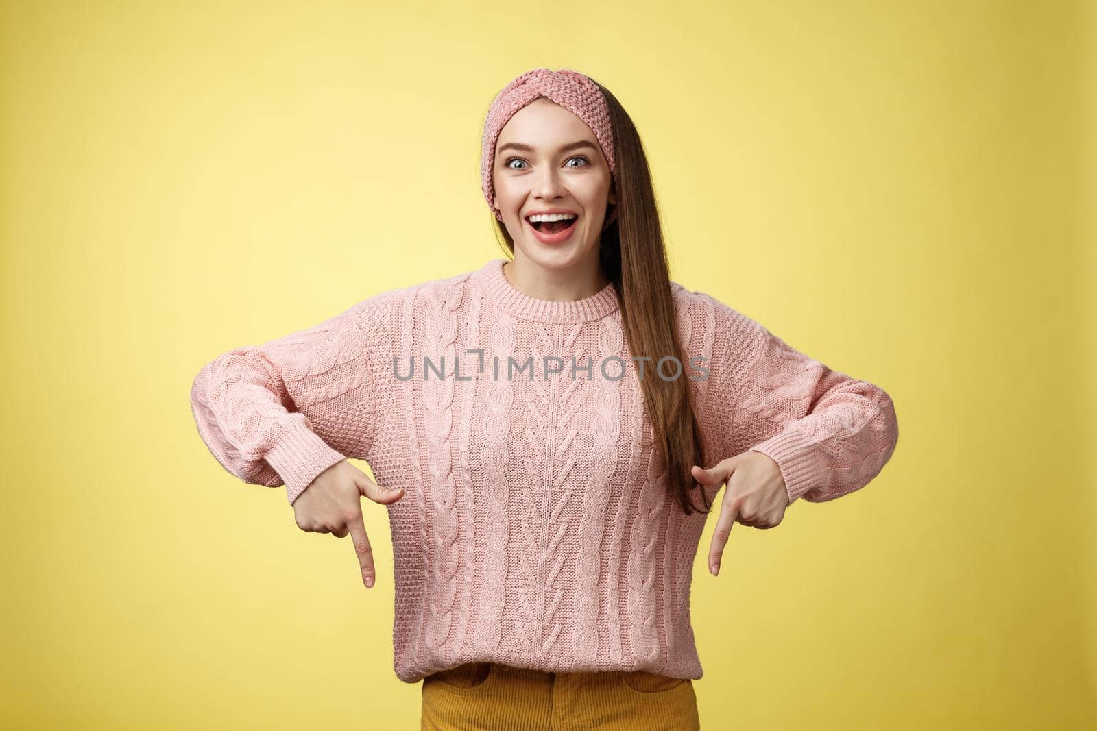 Euphoric attractive young trendy girl in knitted band wearing sweater grinning excited enthusiastic pointing down thrilled of awesome promotion standing amused and overwhelmed over yellow background by Benzoix
